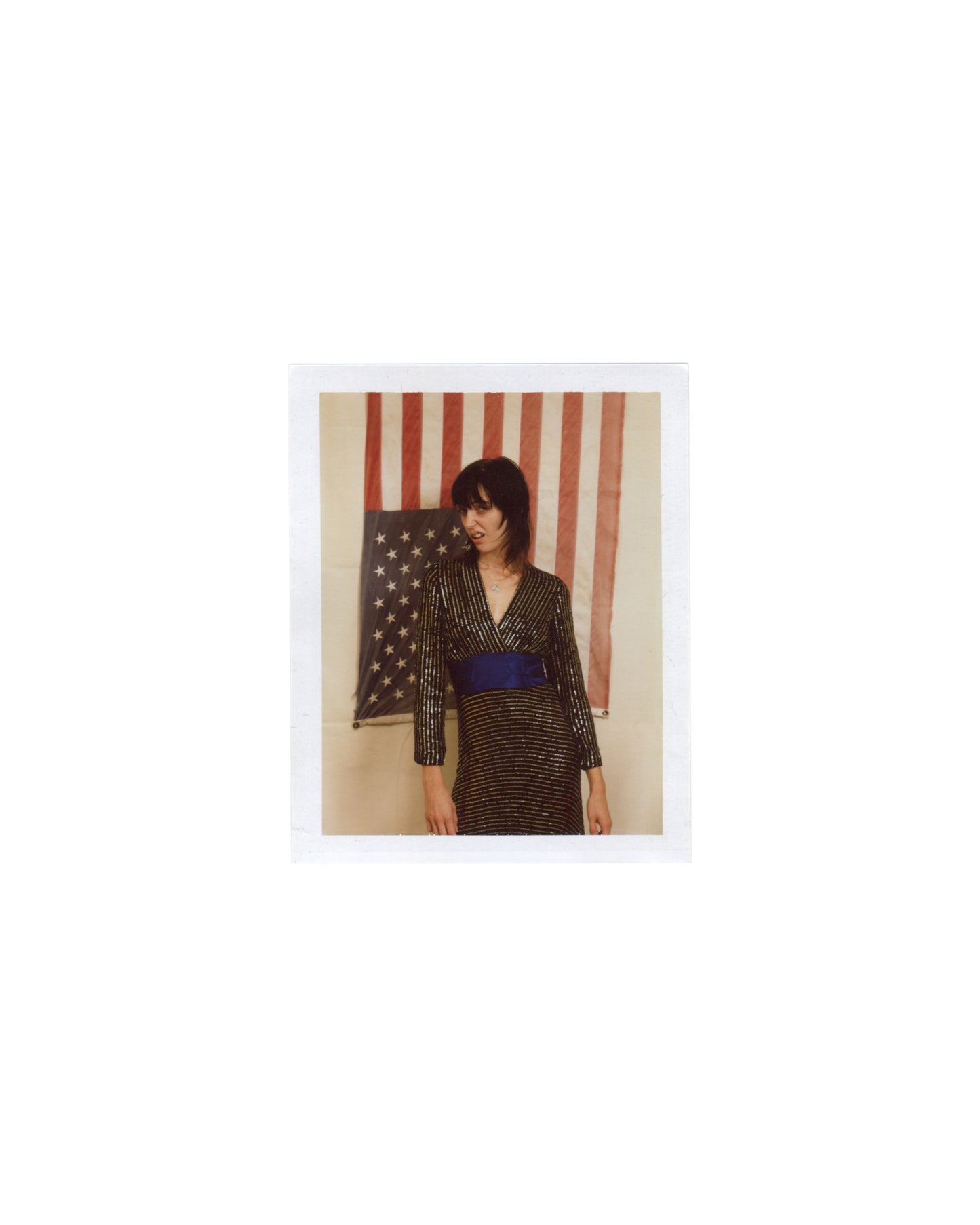 Original Polaroid photograph, Imitation of Christ: A Retrospective, New York, 2002. Woman with dark fringe in gold and black striped wrap dress with blue waist sash, standing before a partial American flag; warm interior light; white Polaroid border.