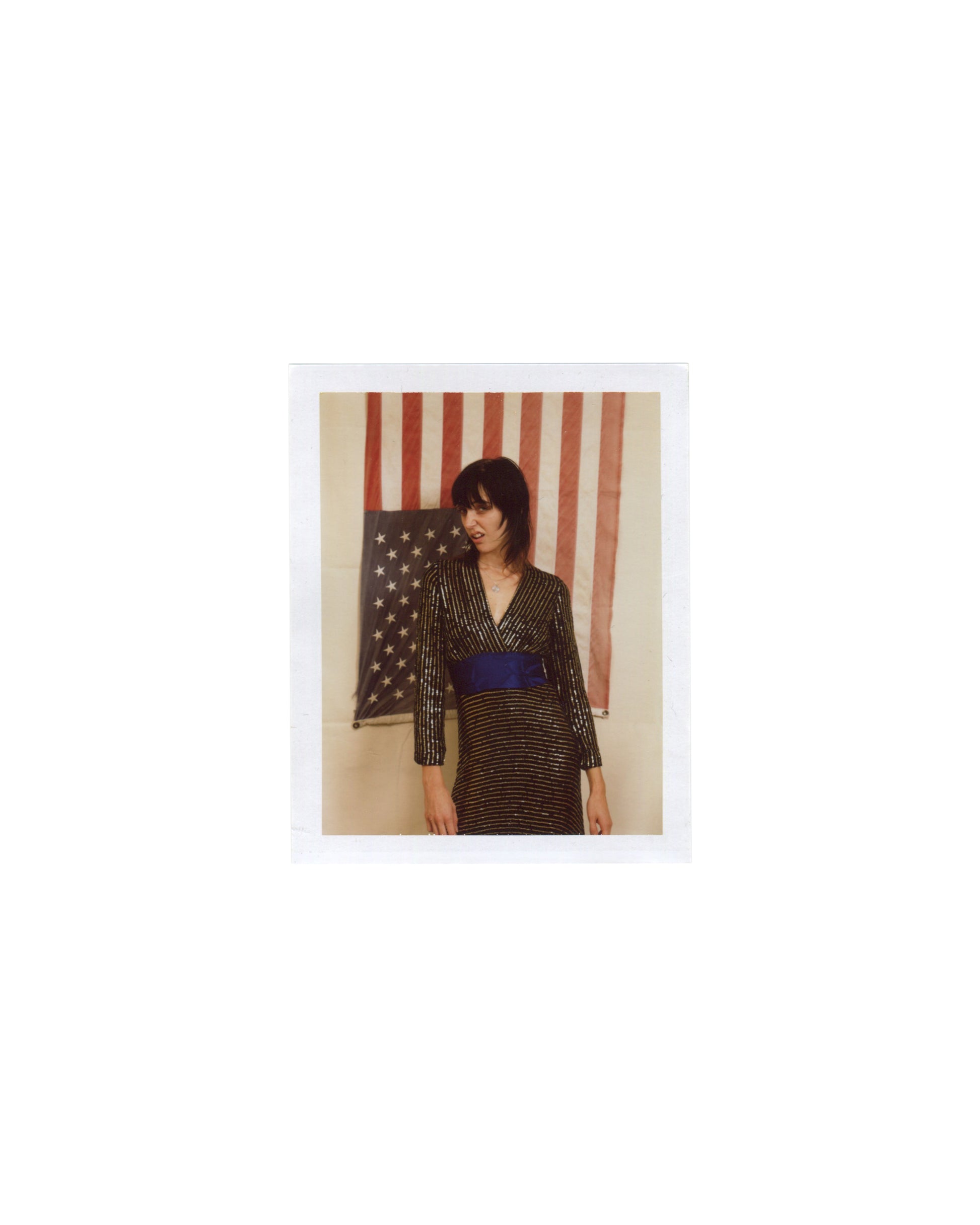 Original Polaroid photograph, Imitation of Christ: A Retrospective, New York, 2002. Woman with dark fringe in gold and black striped wrap dress with blue waist sash, standing before a partial American flag; warm interior light; white Polaroid border.