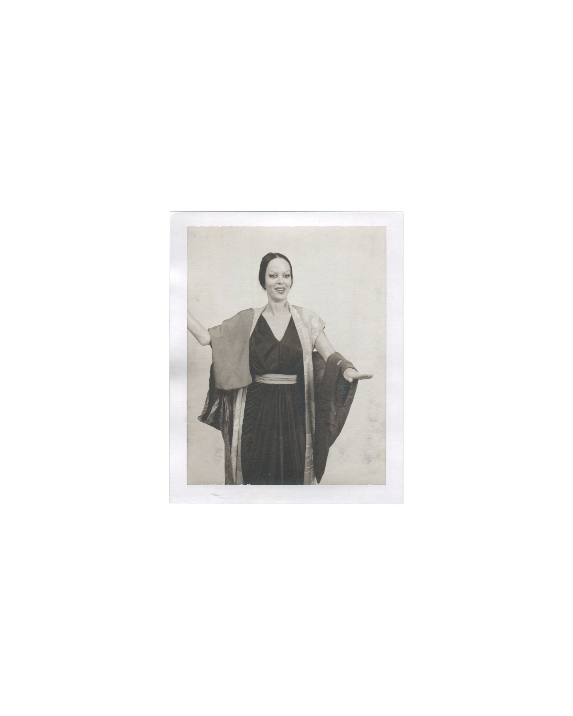 Original Polaroid photograph, Imitation of Christ: A Retrospective, New York, 2002. Black-and-white image of woman in black halter dress and wide-sleeved kimono jacket, arms extended, smiling; neutral background; vintage photographic quality.