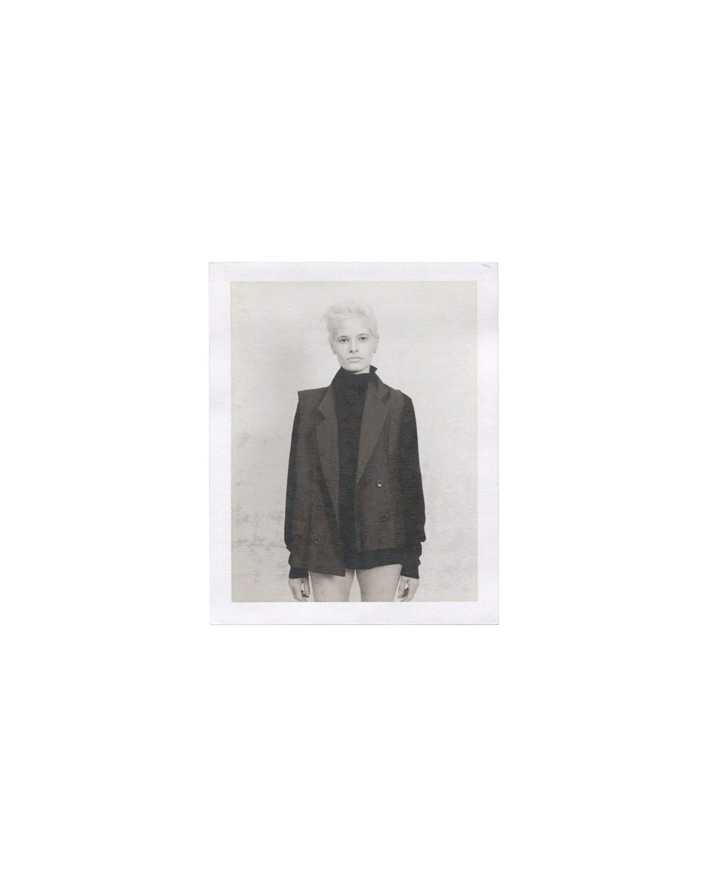 Original Polaroid photograph, Imitation of Christ: A Retrospective, New York, 2002. Black-and-white image of woman with bleached cropped hair in oversized black blazer and turtleneck, direct gaze; grey background; white Polaroid border.