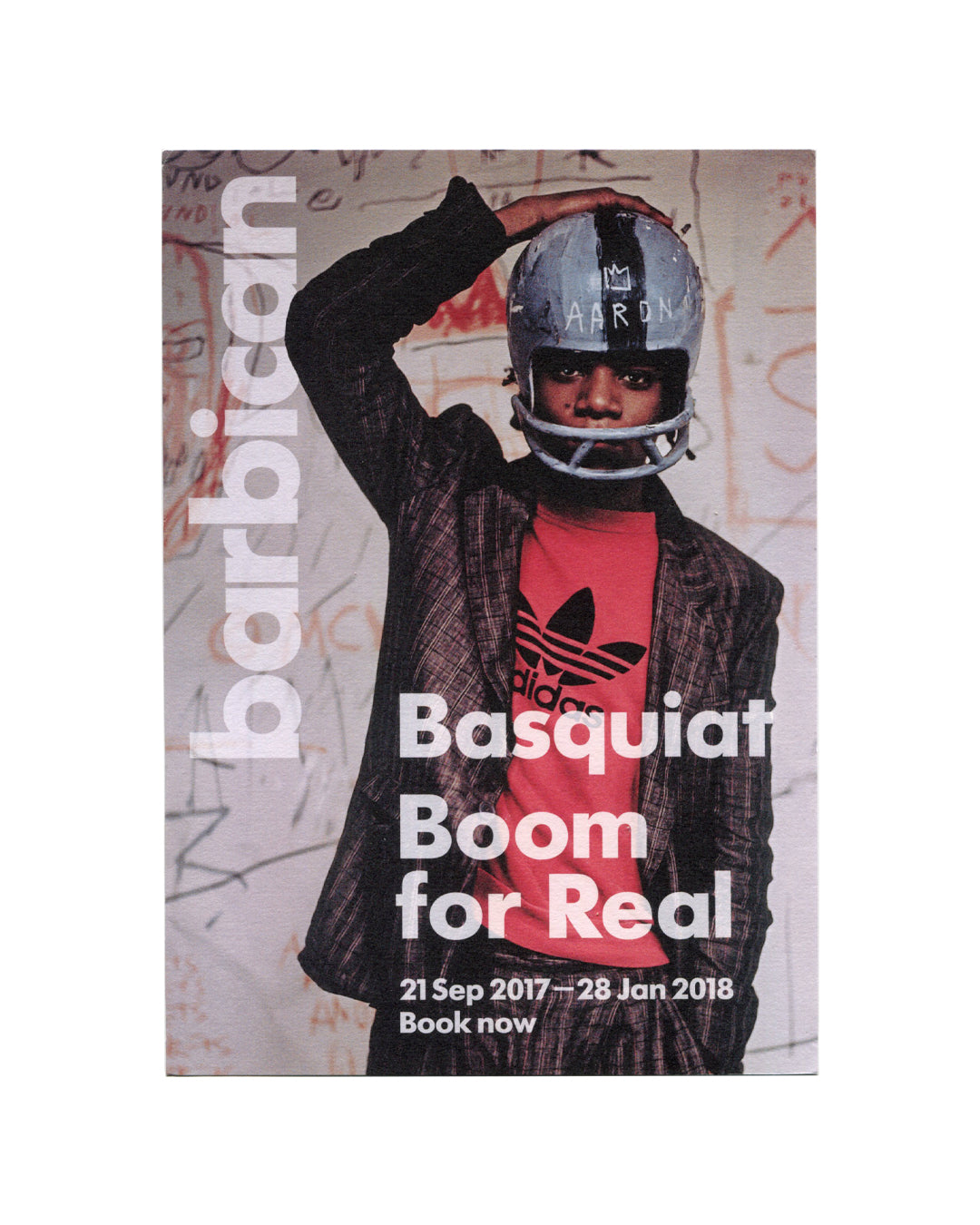 Promotional card recto, Jean-Michel Basquiat: Boom for Real, Barbican Art Gallery, London, 2017. Offset colour on card stock, 21 × 15 cm. Full-bleed photograph by Edo Bertoglio — Basquiat in grey American football helmet marked 'AARON' in white paint, red Adidas T-shirt, open dark plaid jacket; hand raised to helmet; graffiti-covered white wall behind. Left edge: large vertical white sans-serif 'barbican' logotype. Lower centre: bold white sans-serif — 'Basquiat / Boom / for Real / 21 Sep 2017—28 Jan 2018 /