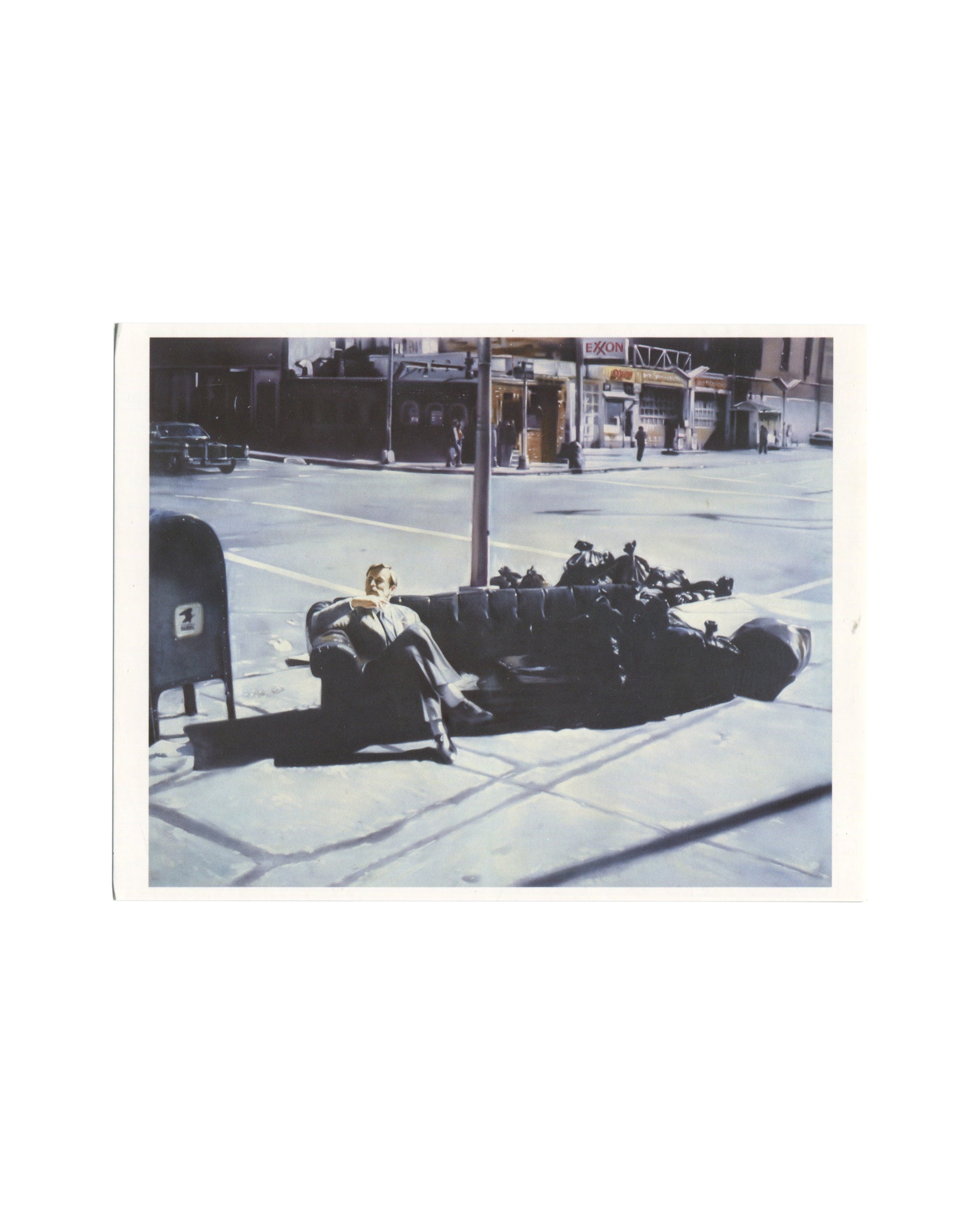 Original 2006 Edition5 artist postcard reproducing Martin Kippenberger's painting Ohne Titel, 1981, from the Lieber Maler, male mir series, showing a colour photorealist painting of a suited man reclining on a dark upholstered sofa abandoned on a city pavement at a wide intersection, a lamp post rising behind him, a US postal mailbox at left, and an Exxon petrol station sign visible across the street in the background, the scene rendered with the flat commercial precision characteristic of the series.