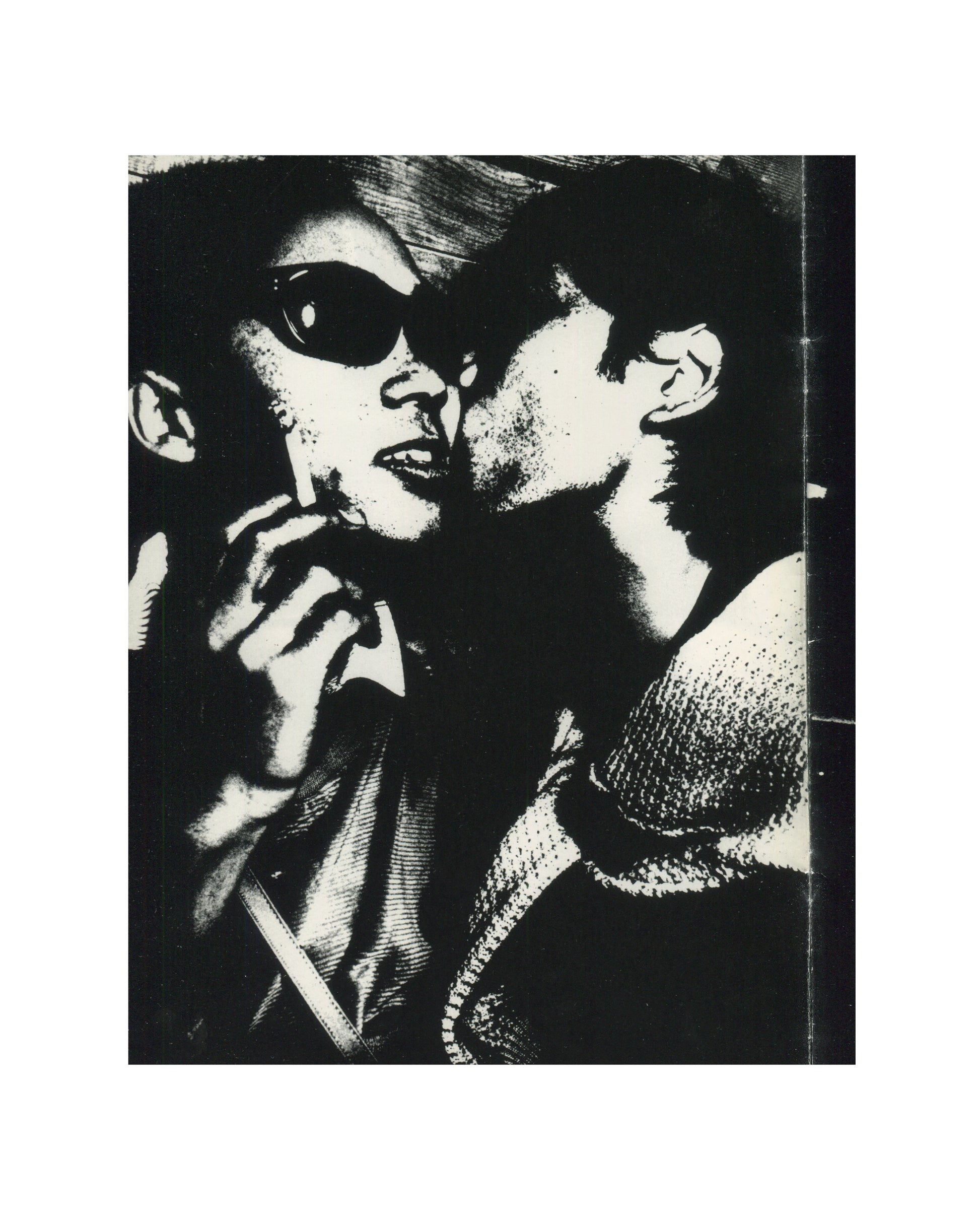 `Keizō Kitajima — Photo Express Tokyo No. 2, February 1979 — interior plate. High-contrast black and white photograph of two figures in extreme close-up: a woman wearing large dark sunglasses holding a cigarette, mouth slightly open, face turned toward a second figure whose lips are close to her cheek. Grain pushed to near-abstraction. Tokyo nightlife, 1979.`