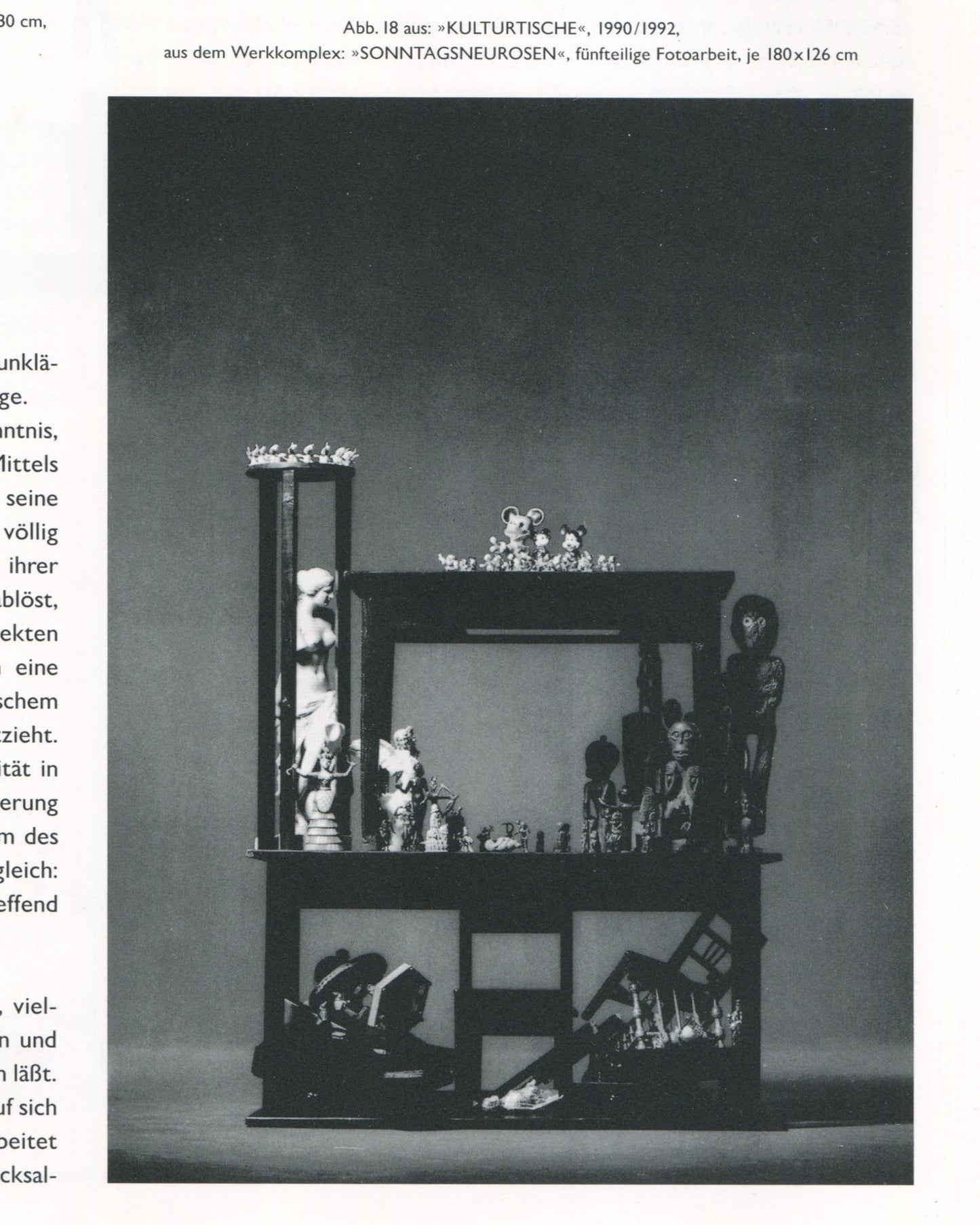 Interior page from Jürgen Klauke Prosecuritas catalogue, 1994. Full-page black-and-white photograph captioned 'Abb. 18 aus: »KULTURTISCHE«, 1990/1992, aus dem Werkkomplex: »SONNTAGSNEUROSEN«, fünfteilige Fotoarbeit, je 180×126 cm'. A two-storey vitrine-like wooden structure filled with figurines, tribal objects, a classical nude sculpture, Mickey Mouse toys, and chess pieces arranged across its shelves and floor.