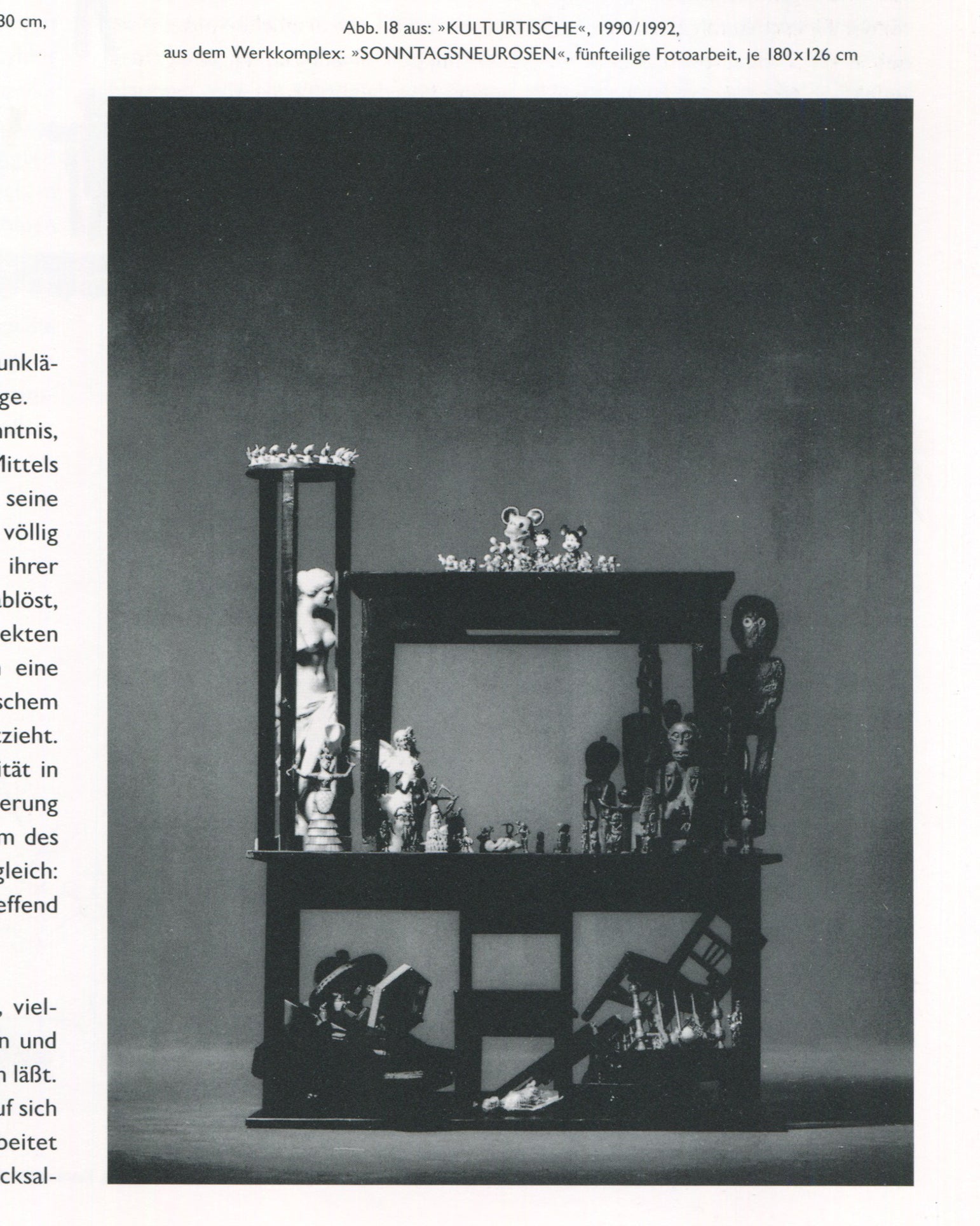 Interior page from Jürgen Klauke Prosecuritas catalogue, 1994. Full-page black-and-white photograph captioned 'Abb. 18 aus: »KULTURTISCHE«, 1990/1992, aus dem Werkkomplex: »SONNTAGSNEUROSEN«, fünfteilige Fotoarbeit, je 180×126 cm'. A two-storey vitrine-like wooden structure filled with figurines, tribal objects, a classical nude sculpture, Mickey Mouse toys, and chess pieces arranged across its shelves and floor.