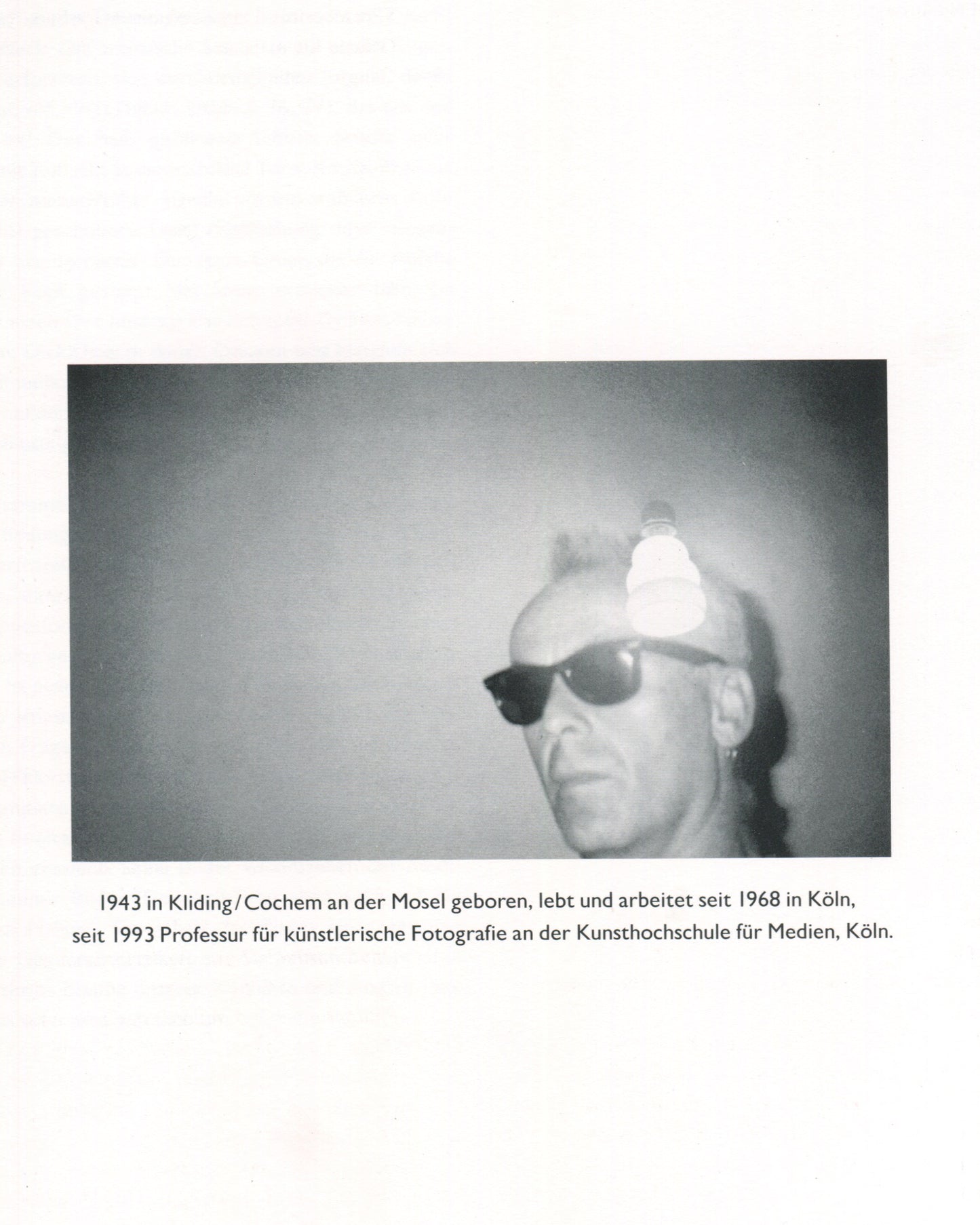 Interior page from Jürgen Klauke Prosecuritas catalogue, 1994. Black-and-white photograph of Jürgen Klauke from the shoulders up, wearing dark sunglasses and a small earring, with a bright light source flaring above his head against a plain grey ground. Below the image, biographical text in German: '1943 in Kliding/Cochem an der Mosel geboren, lebt und arbeitet seit 1968 in Köln, seit 1993 Professur für künstlerische Fotografie an der Kunsthochschule für Medien, Köln.