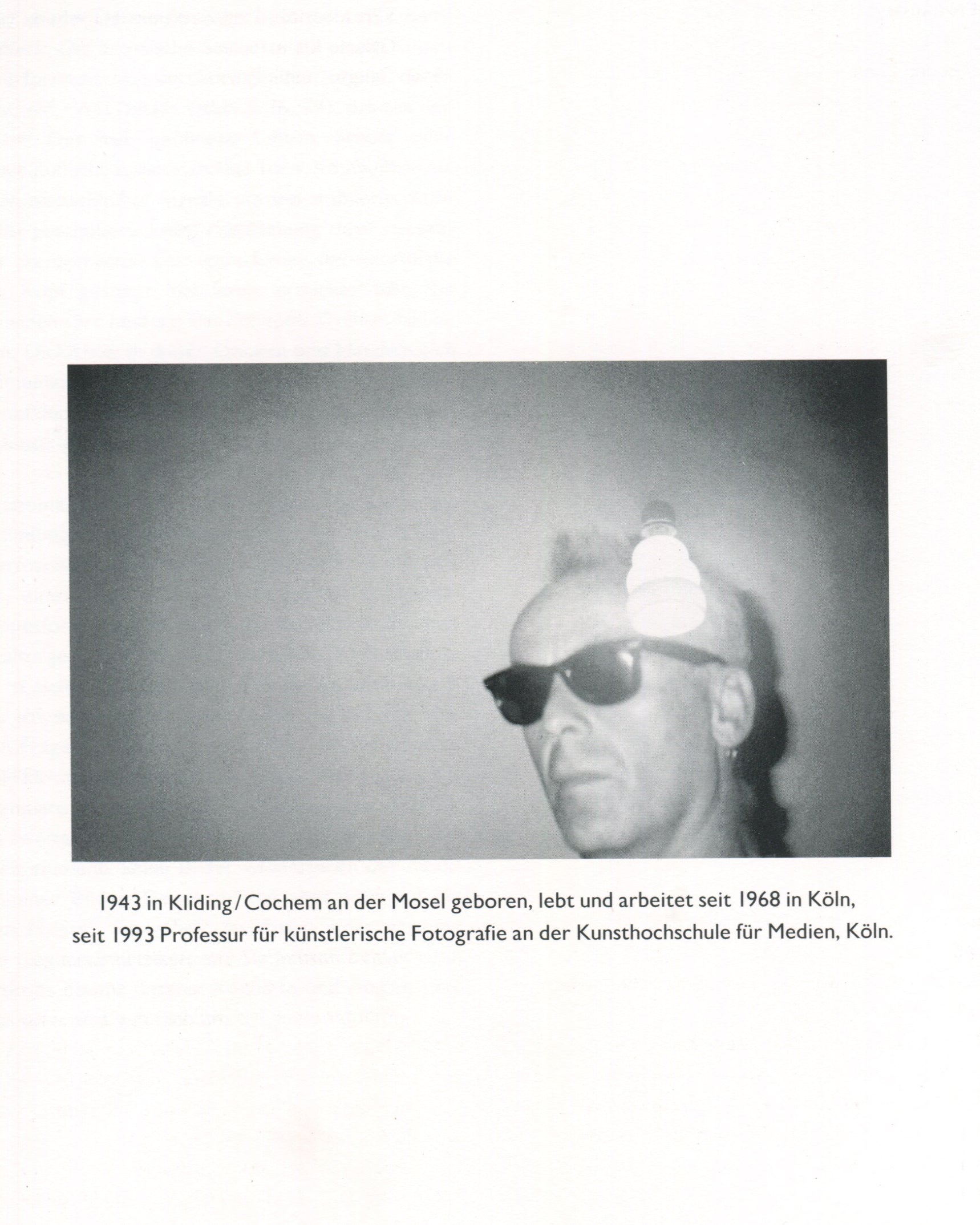 Interior page from Jürgen Klauke Prosecuritas catalogue, 1994. Black-and-white photograph of Jürgen Klauke from the shoulders up, wearing dark sunglasses and a small earring, with a bright light source flaring above his head against a plain grey ground. Below the image, biographical text in German: '1943 in Kliding/Cochem an der Mosel geboren, lebt und arbeitet seit 1968 in Köln, seit 1993 Professur für künstlerische Fotografie an der Kunsthochschule für Medien, Köln.