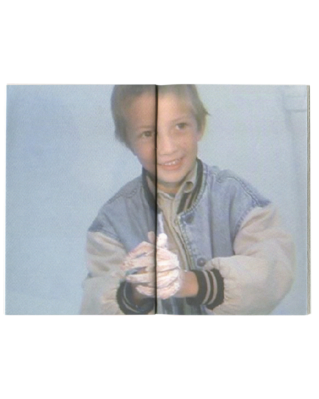 Terence Koh, The Whole Family, interior full-bleed spread. Colour video-still photograph of a young boy in a denim and grey varsity jacket, smiling, hands raised and crossed at wrist, against a pale blue-grey background. Low-resolution, VHS-quality image. Peres Projects, Los Angeles, 2008.