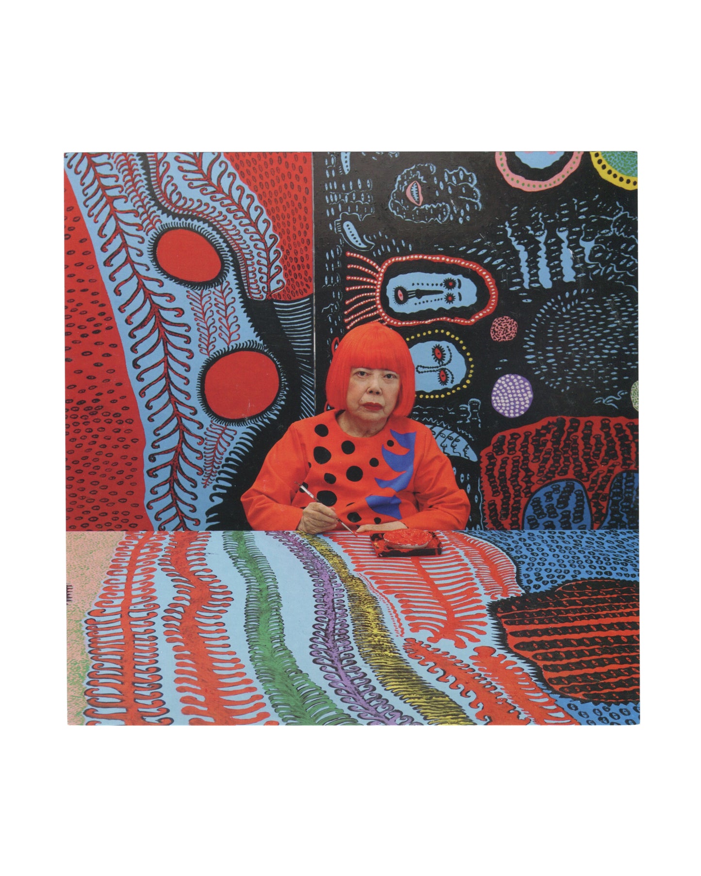 2017 Yayoi Kusama exhibition invitation card featuring a studio portrait of the artist seated at a table surrounded by vividly colored *My Eternal Soul* paintings. Kusama wears her signature bright red wig and an orange dress with black polka dots, visually blending into the dense field of reds, blues, dots, and biomorphic patterns surrounding her.