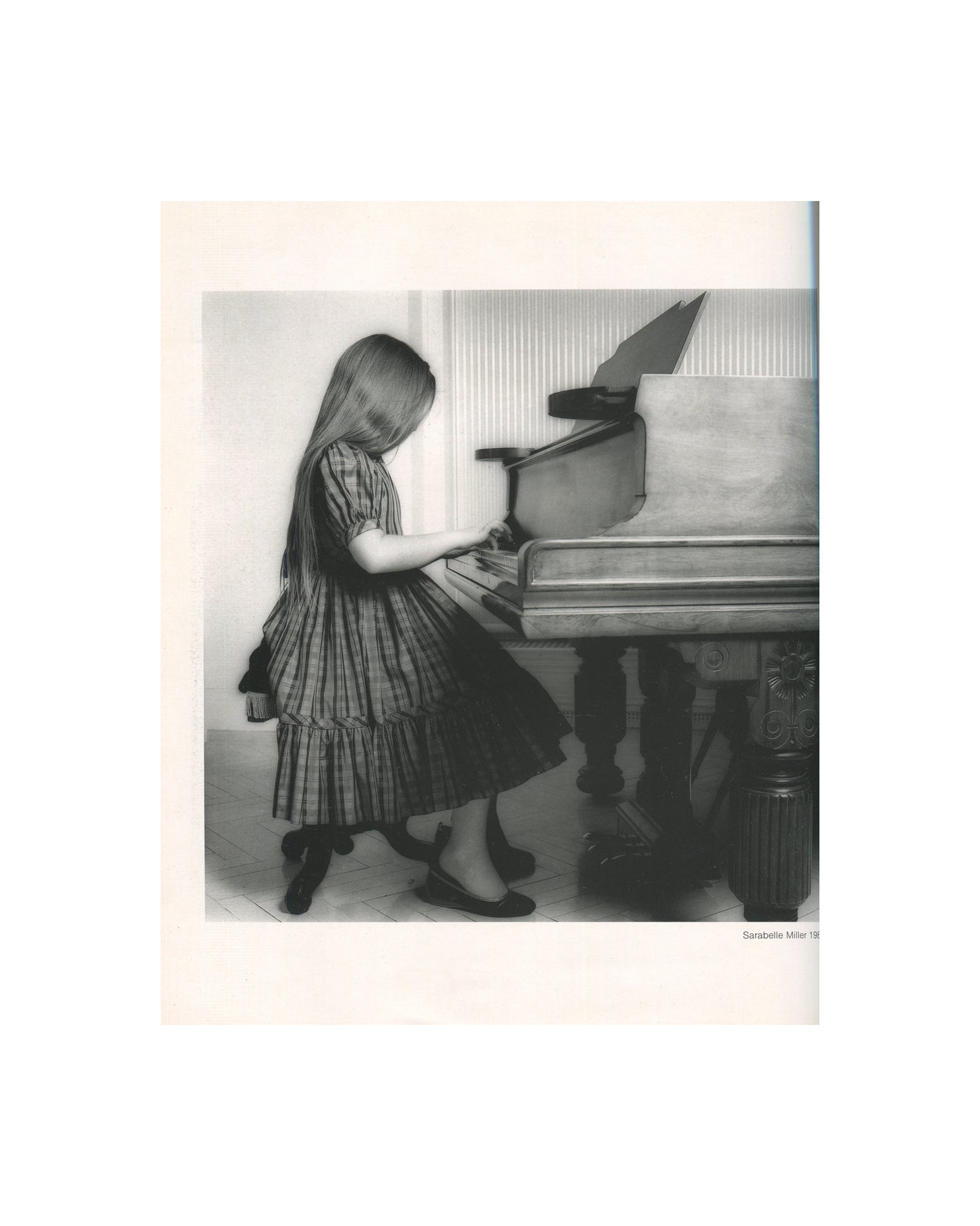 Robert Mapplethorpe — Fotografie, Idea Books Edizioni, Milan, 1983. Interior plate: Sarabelle Miller, 1981. Black-and-white photograph of a young girl with long hair in a striped dress and black shoes, standing at a grand piano, pressing the keys, head bowed. Sheet music open on the stand. Caption lower right: 'Sarabelle Miller 19[81]'. Photography monograph, Germano Celant.