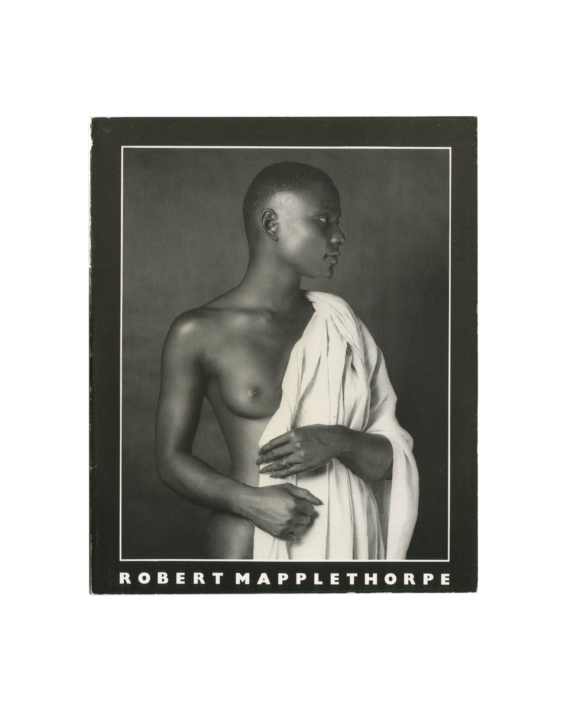 Robert Mapplethorpe — Fotografie, Idea Books Edizioni, Milan, 1983. Back cover. Black ground with white border rule. Black-and-white photograph: a young Black woman in profile, nude to the waist, holding a white draped cloth across her body, rings on her fingers, eyes downcast. Title in white block capitals along lower edge: 'ROBERT MAPPLETHORPE'. Photography monograph, Italian edition.