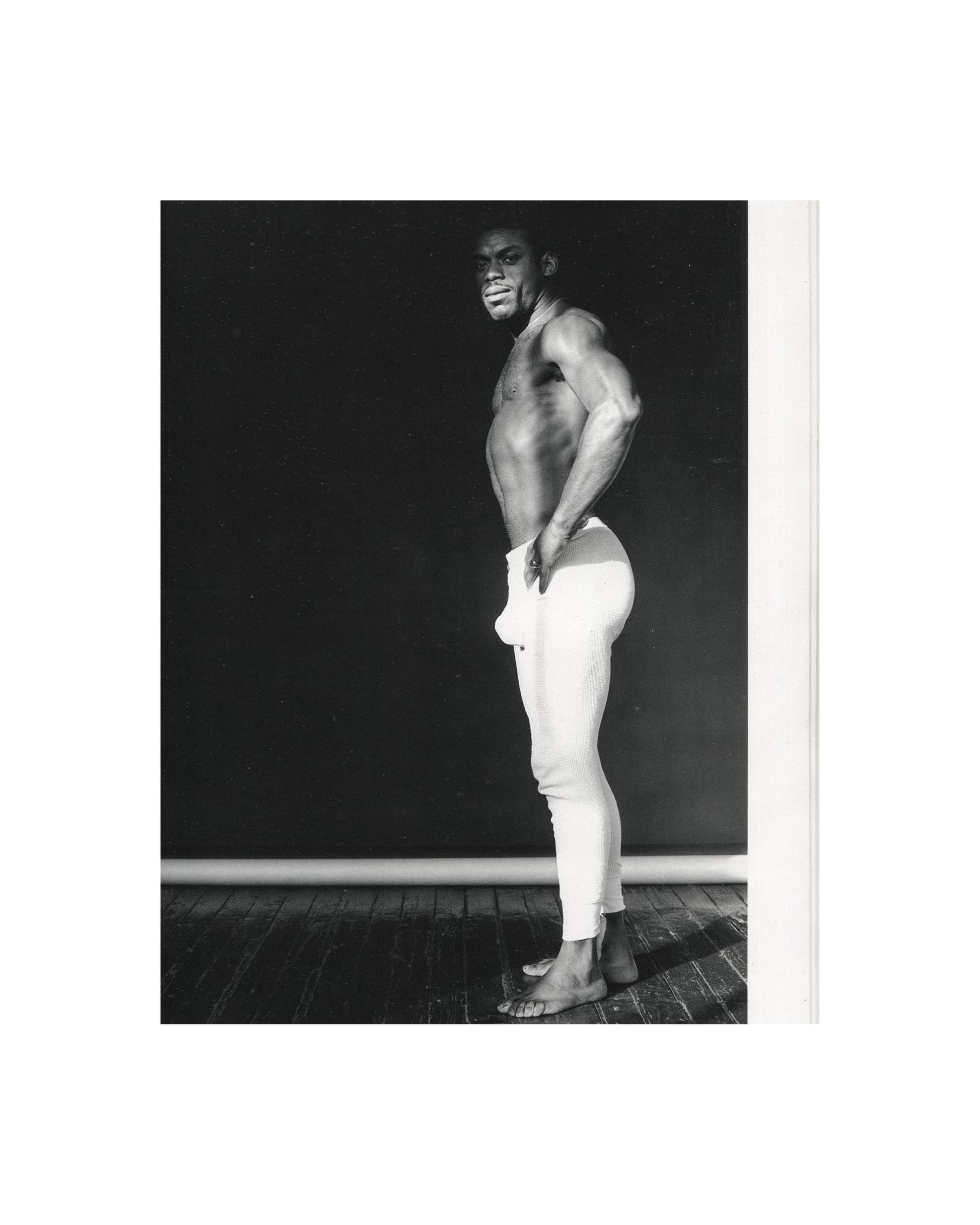 Robert Mapplethorpe — Fotografie, Idea Books Edizioni, Milan, 1983. Interior plate: black-and-white studio photograph. A Black man, bare-chested and muscular, wearing white trousers pushed down at the back, standing in three-quarter profile, looking back over his shoulder toward the camera, smiling. Dark wall and wooden floor visible. Classical Mapplethorpe figure study. Photography monograph, Germano Celant, The New Rare.