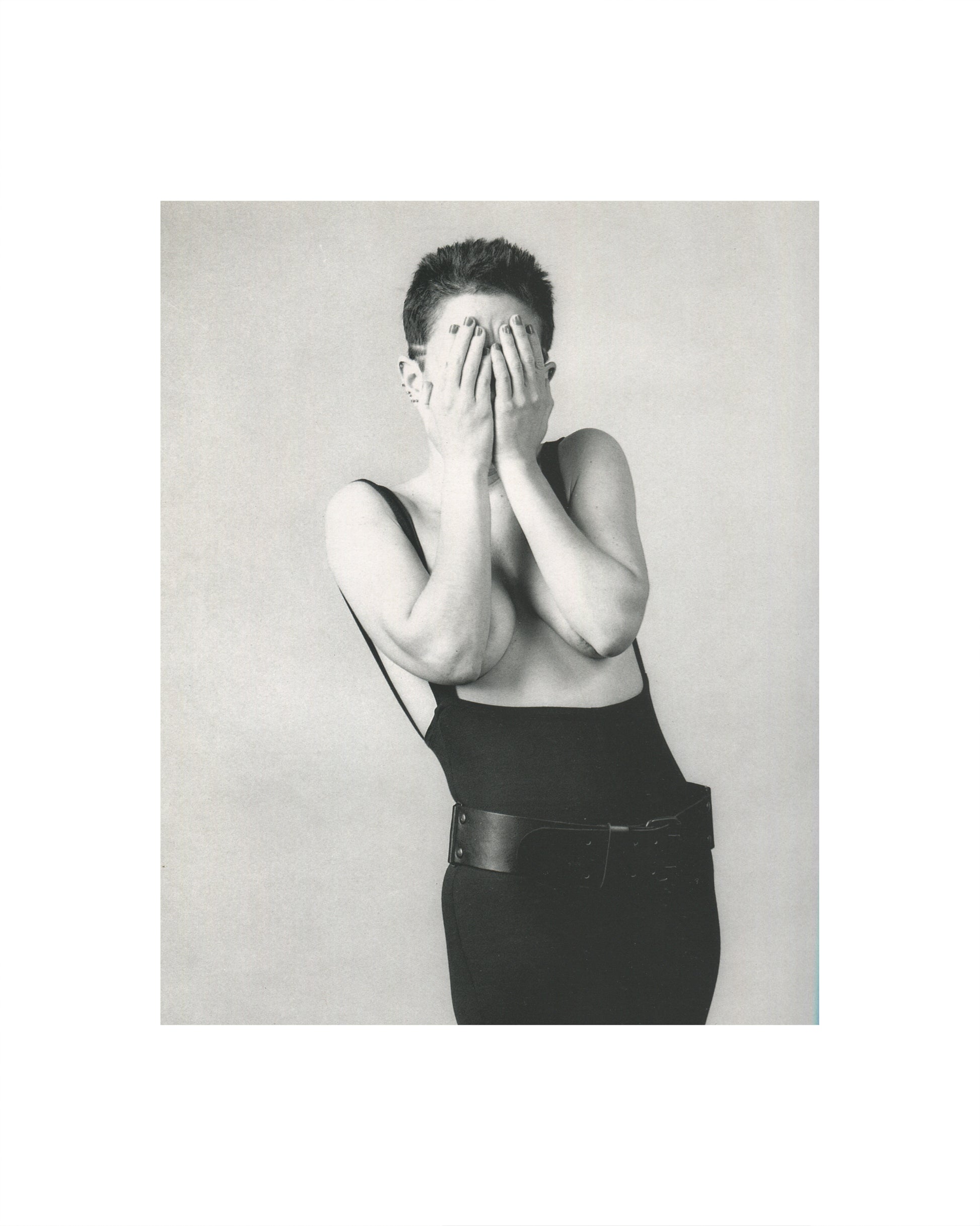 Robert Mapplethorpe — Fotografie, Idea Books Edizioni, Milan, 1983. Interior plate: black-and-white studio photograph. A figure with short dark hair, bare-shouldered, wearing black dungarees and a wide leather belt, both hands raised to cover their face, painted fingernails visible, small earring. Neutral grey ground. High-contrast Mapplethorpe studio lighting. Photography monograph, Germano Celant, The New Rare.