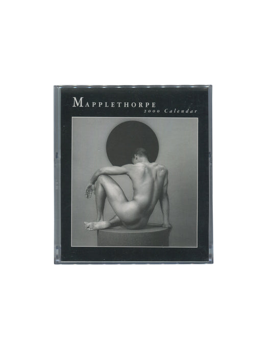 Cover insert of Robert Mapplethorpe, 2000 Calendar, CD case edition, teNeues, 1999, showing the calendar housed in a clear plastic CD jewel case, with a black printed insert reading MAPPLETHORPE / 2000 Calendar in white serif type at upper left, and a black-and-white photograph of a nude male figure seated on a plinth with his back to the camera, knees drawn up, against a circular dark disc.