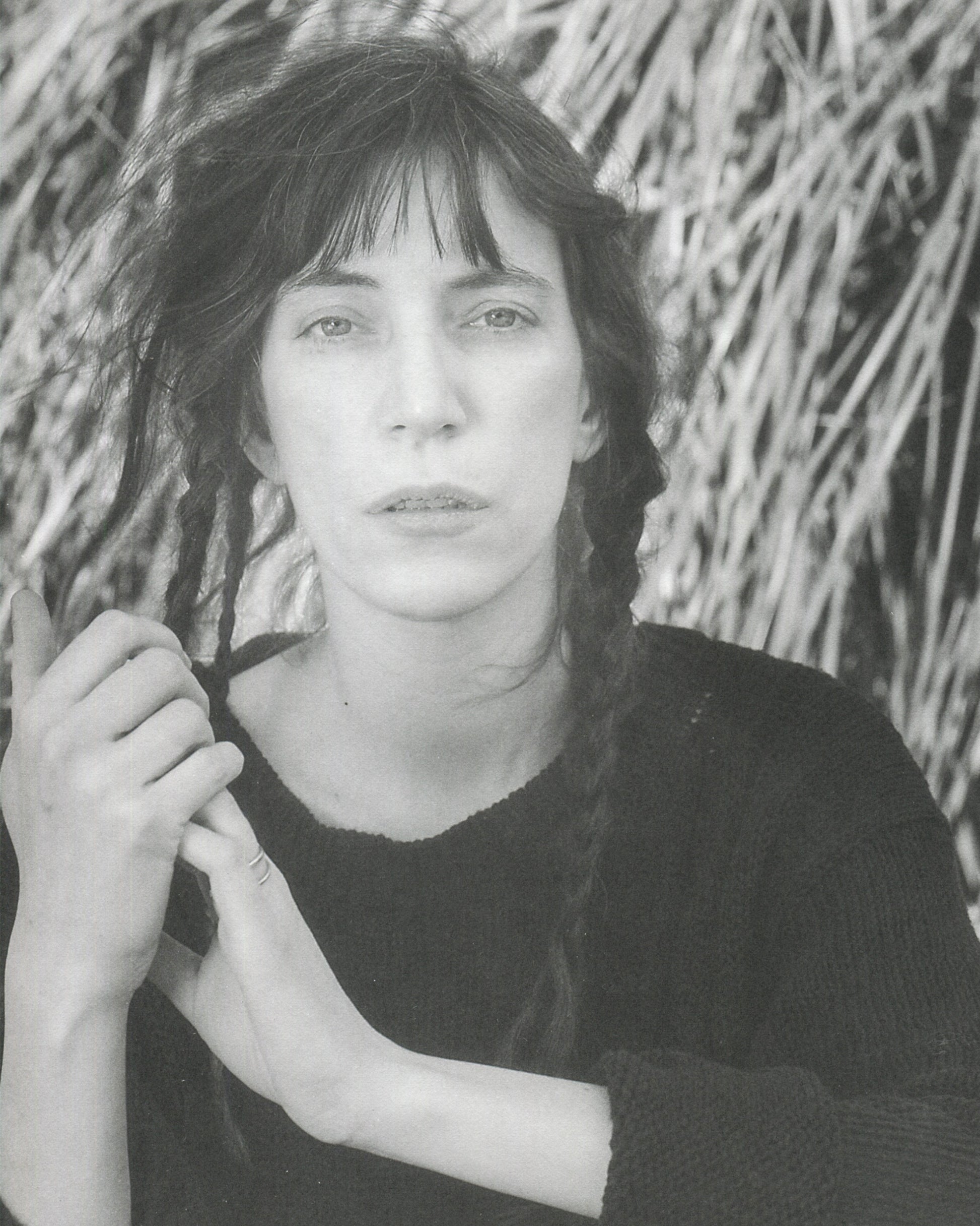Interior page from Robert Mapplethorpe, Some Women, Bulfinch Press, 1992, showing a black-and-white portrait of Patti Smith, photographed outdoors against a background of dried straw or reeds, wearing a dark knitted sweater, her dark hair loose with a fringe, holding a braid of her hair in both hands, looking directly at the camera with a composed, direct expression.