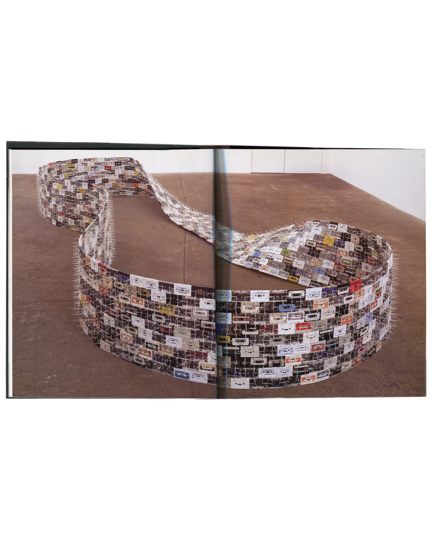 Interior spread from Christian Marclay, Phaidon Contemporary Artists Series, Phaidon Press, 2005, showing a full-bleed colour photograph across both pages of a large floor-based installation comprising hundreds of cassette tapes woven together with wire or thread into a long, wide, undulating ribbon that coils and loops across a wooden gallery floor, the cassette labels visible in white, red, blue, and multicolour against the pale tape bodies.