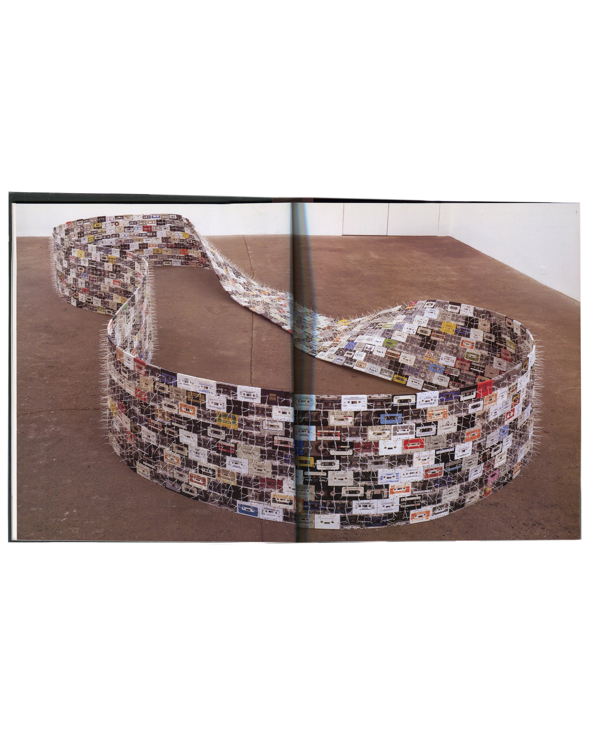 Interior spread from Christian Marclay, Phaidon Contemporary Artists Series, Phaidon Press, 2005, showing a full-bleed colour photograph across both pages of a large floor-based installation comprising hundreds of cassette tapes woven together with wire or thread into a long, wide, undulating ribbon that coils and loops across a wooden gallery floor, the cassette labels visible in white, red, blue, and multicolour against the pale tape bodies.