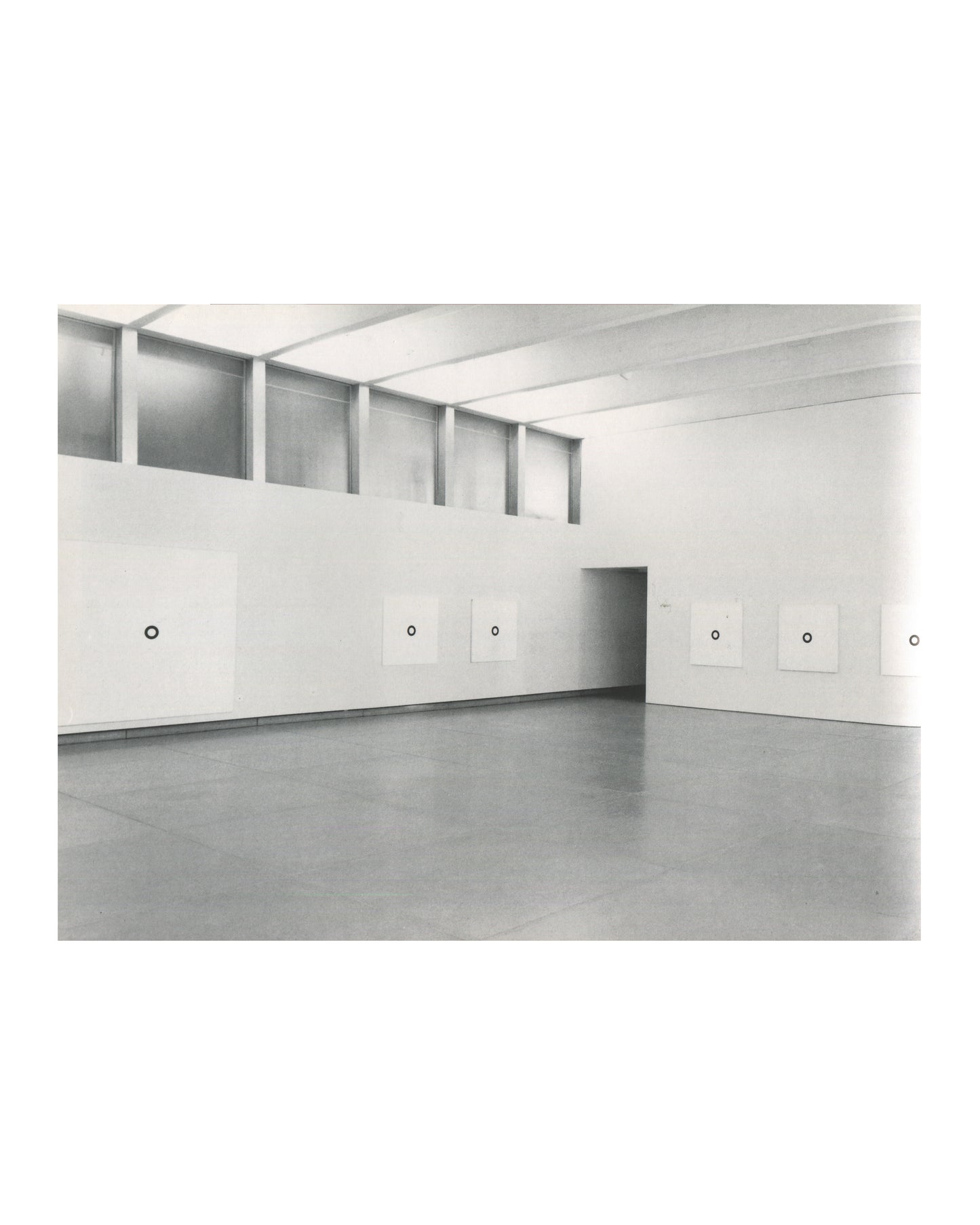 Interior page, Olivier Mosset, Verlag Lars Müller, Baden, 1990. Black-and-white installation photograph — large gallery with clerestory windows; six white square canvases each with a single small black circle hung at regular intervals; grey concrete floor. Very good condition.