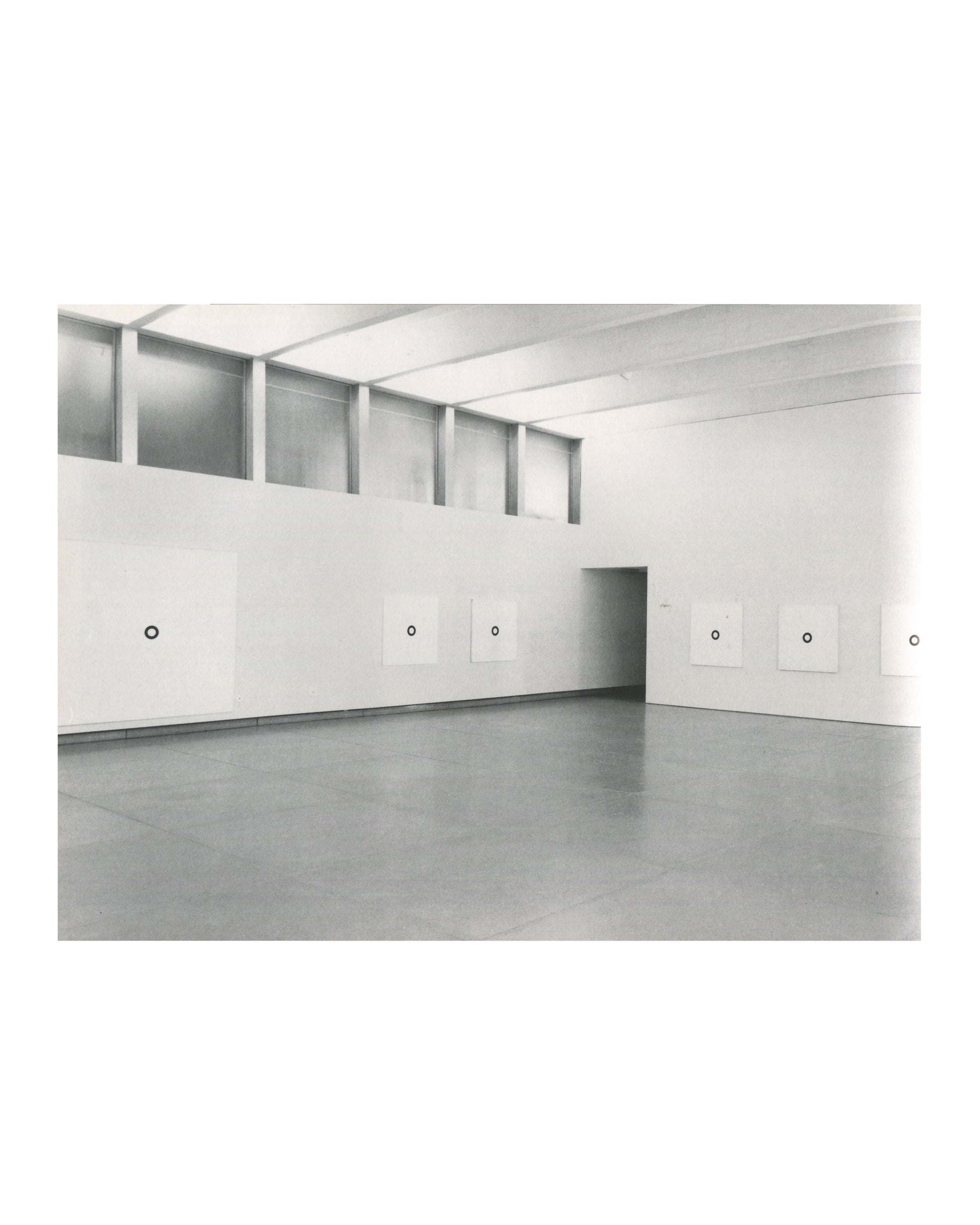 Interior page, Olivier Mosset, Verlag Lars Müller, Baden, 1990. Black-and-white installation photograph — large gallery with clerestory windows; six white square canvases each with a single small black circle hung at regular intervals; grey concrete floor. Very good condition.