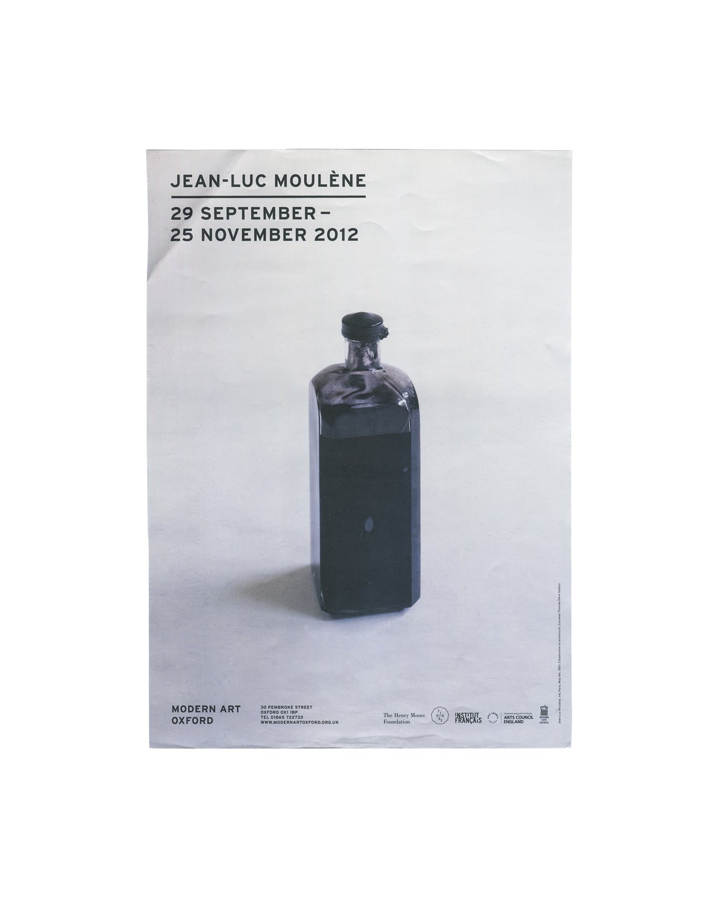 Original exhibition poster, Jean-Luc Moulène, Modern Art Oxford, 29 September – 25 November 2012. Offset lithograph on paper, 29.5 × 42 cm. White ground. Upper left: bold black sans-serif text 'JEAN-LUC MOULÈNE' with exhibition dates '29 SEPTEMBER – / 25 NOVEMBER 2012' below. Centre: large colour photograph of Anti Paris, May 6th, 2001 — a dark opaque glass bottle with a black stopper, isolated against a pale neutral ground, casting a soft shadow. Lower left: 'MODERN ART OXFORD' in bold black sans-serif wit