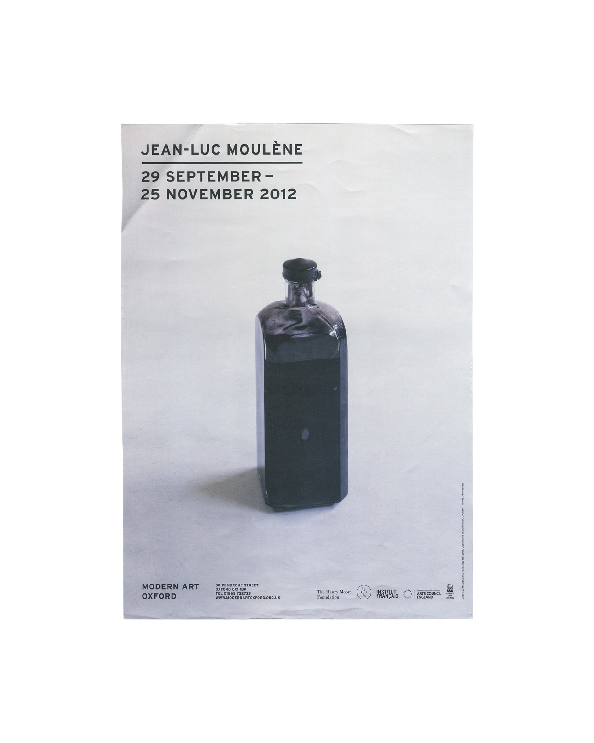 Original exhibition poster, Jean-Luc Moulène, Modern Art Oxford, 29 September – 25 November 2012. Offset lithograph on paper, 29.5 × 42 cm. White ground. Upper left: bold black sans-serif text 'JEAN-LUC MOULÈNE' with exhibition dates '29 SEPTEMBER – / 25 NOVEMBER 2012' below. Centre: large colour photograph of Anti Paris, May 6th, 2001 — a dark opaque glass bottle with a black stopper, isolated against a pale neutral ground, casting a soft shadow. Lower left: 'MODERN ART OXFORD' in bold black sans-serif wit