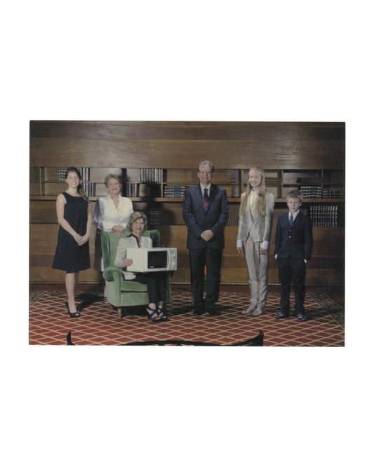 Original 2012 The Virgins Show Family Business New York exhibition invitation card, recto showing a colour photograph of six figures posed in a wood-panelled library interior with floor-to-ceiling bookshelves: from left, a young woman in a black dress, an older woman in white, a seated woman in a green armchair holding a microwave oven on her lap, a man in a dark suit, a teenage girl in a beige suit, and a young boy in a dark suit, standing on a red geometric-patterned carpet