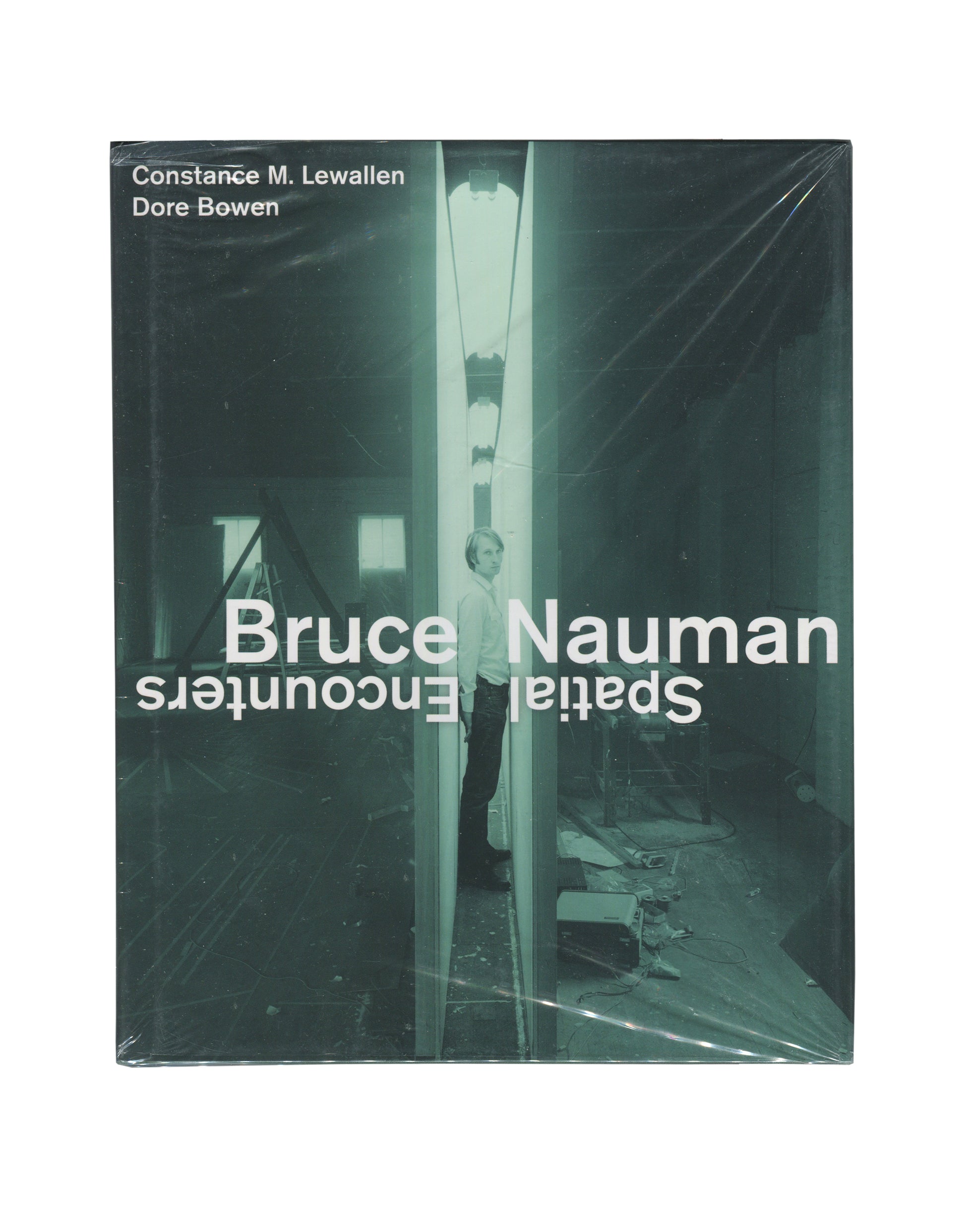 Monograph front cover, Bruce Nauman: Spatial Encounters, Constance M. Lewallen and Dore Bowen, University of California Press, 2019. Sealed in distributor shrinkwrap. Dark teal-green ground. Full-bleed black-and-white photograph — Bruce Nauman standing inside a narrow corridor installation, fluorescent light strip overhead, mirror surfaces on either side producing infinite reflection; figure slightly off-centre, looking toward camera. Upper left: white sans-serif — 'Constance M. Lewallen / Dore Bowen'. 