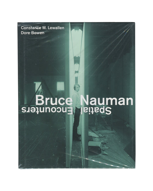 Monograph front cover, Bruce Nauman: Spatial Encounters, Constance M. Lewallen and Dore Bowen, University of California Press, 2019. Sealed in distributor shrinkwrap. Dark teal-green ground. Full-bleed black-and-white photograph — Bruce Nauman standing inside a narrow corridor installation, fluorescent light strip overhead, mirror surfaces on either side producing infinite reflection; figure slightly off-centre, looking toward camera. Upper left: white sans-serif — 'Constance M. Lewallen / Dore Bowen'. 