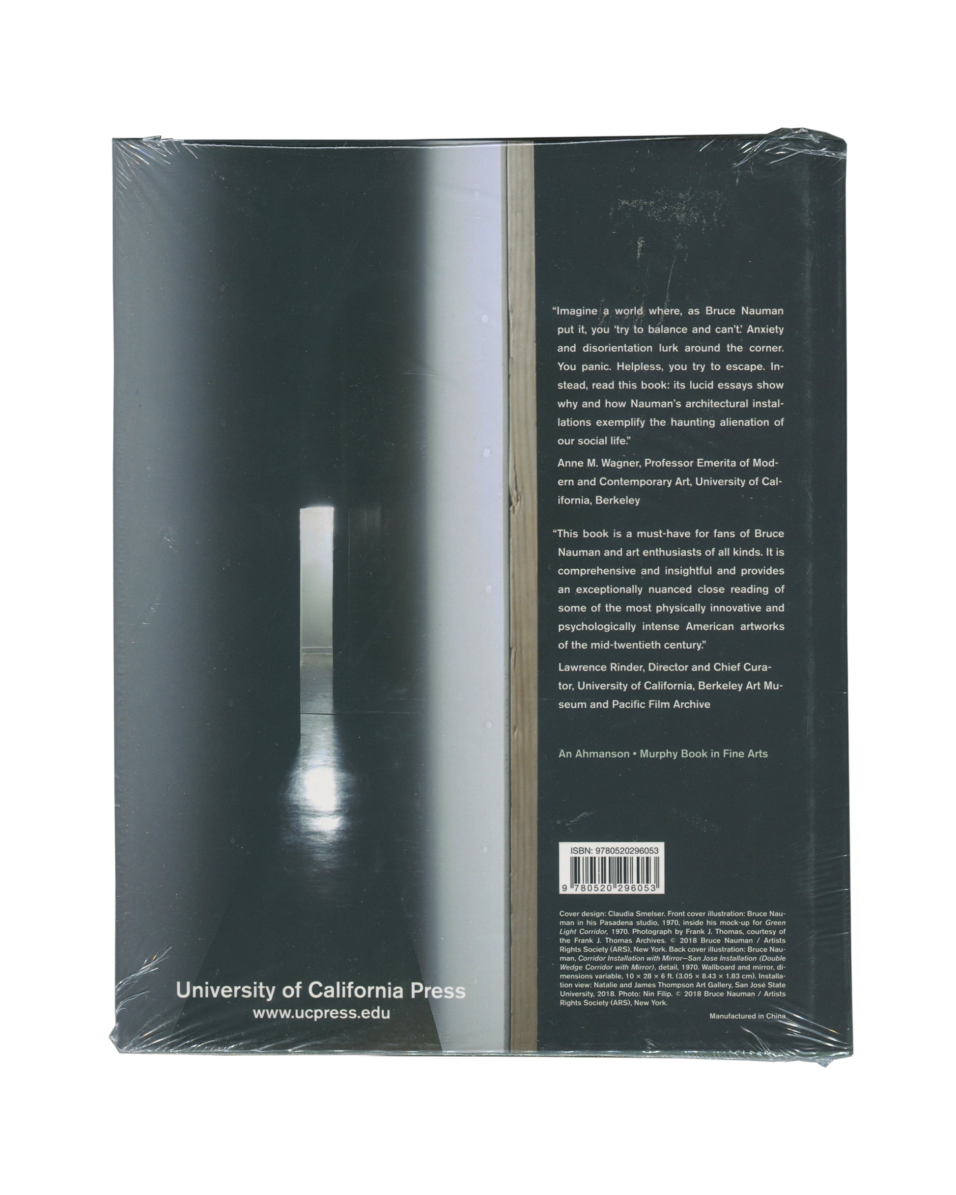 Monograph back cover, Bruce Nauman: Spatial Encounters, University of California Press, 2019. Sealed in distributor shrinkwrap. Dark teal-green ground. Full-bleed photograph — narrow corridor installation, single vertical strip of light at centre, reflective floor, deep spatial recession. Right column: two endorsement quotes — Anne M. Wagner, Professor Emerita of Modern and Contemporary Art, UC Berkeley; Lawrence Rinder, Director and Chief Curator, UC Berkeley Art Museum and Pacific Film Archive. 