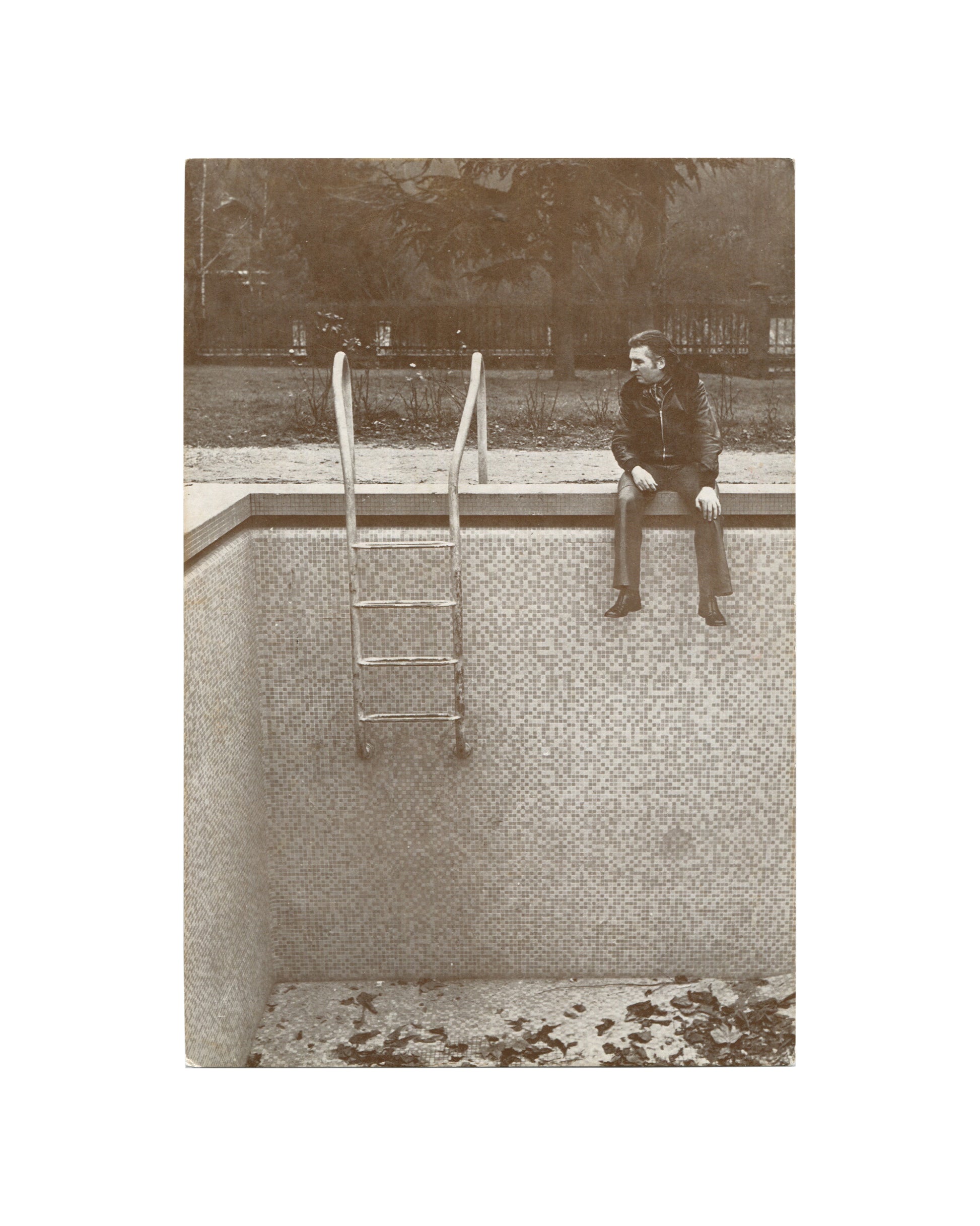  Original 1973 Galerie Stadler exhibition invitation for Francis Naves, À propos des piscines…, Paris, front, sepia-toned photograph of a solitary figure in a dark jacket seated on the edge of an empty tiled swimming pool, legs dangling over the side, looking to the right; a metal ladder descends into the empty pool at left; fallen leaves visible on the pool floor; trees and a fence in the background.