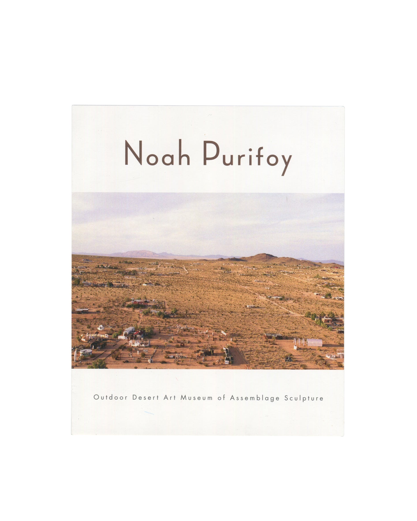 Noah Purifoy Foundation museum brochure, Outdoor Desert Art Museum of Assemblage Sculpture, Joshua Tree, c. 2022, front cover, white ground with Noah Purifoy in large brown serif type at top; below a full-width aerial colour photograph of the desert site showing scattered assemblage sculptures across a flat arid landscape with mountains in the background; Outdoor Desert Art Museum of Assemblage Sculpture in small brown serif type at foot.