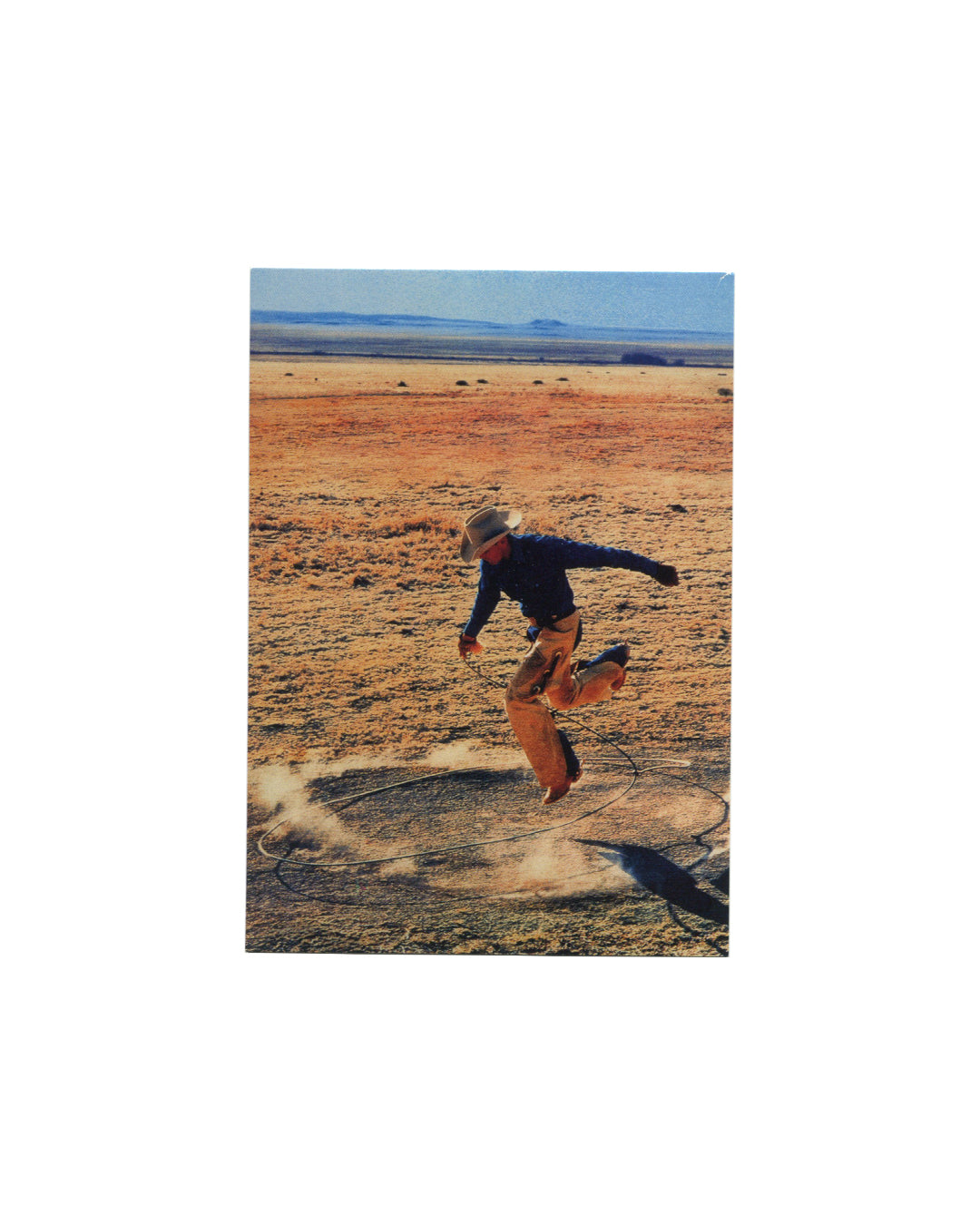 Postcard recto, Richard Prince, Untitled (cowboy), 2003, Solomon R. Guggenheim Museum, New York, 2007. Offset colour reproduction on cream card. Full-bleed vertical image — cowboy in dark navy shirt, tan chaps, and wide-brimmed hat mid-stride across arid flat desert plain; lasso coiled on dusty ground around feet; vast ochre landscape, pale blue sky and distant mesa behind. Ektacolor photograph, edition of 2, 40 × 30 inches. Light handling only, very good condition.