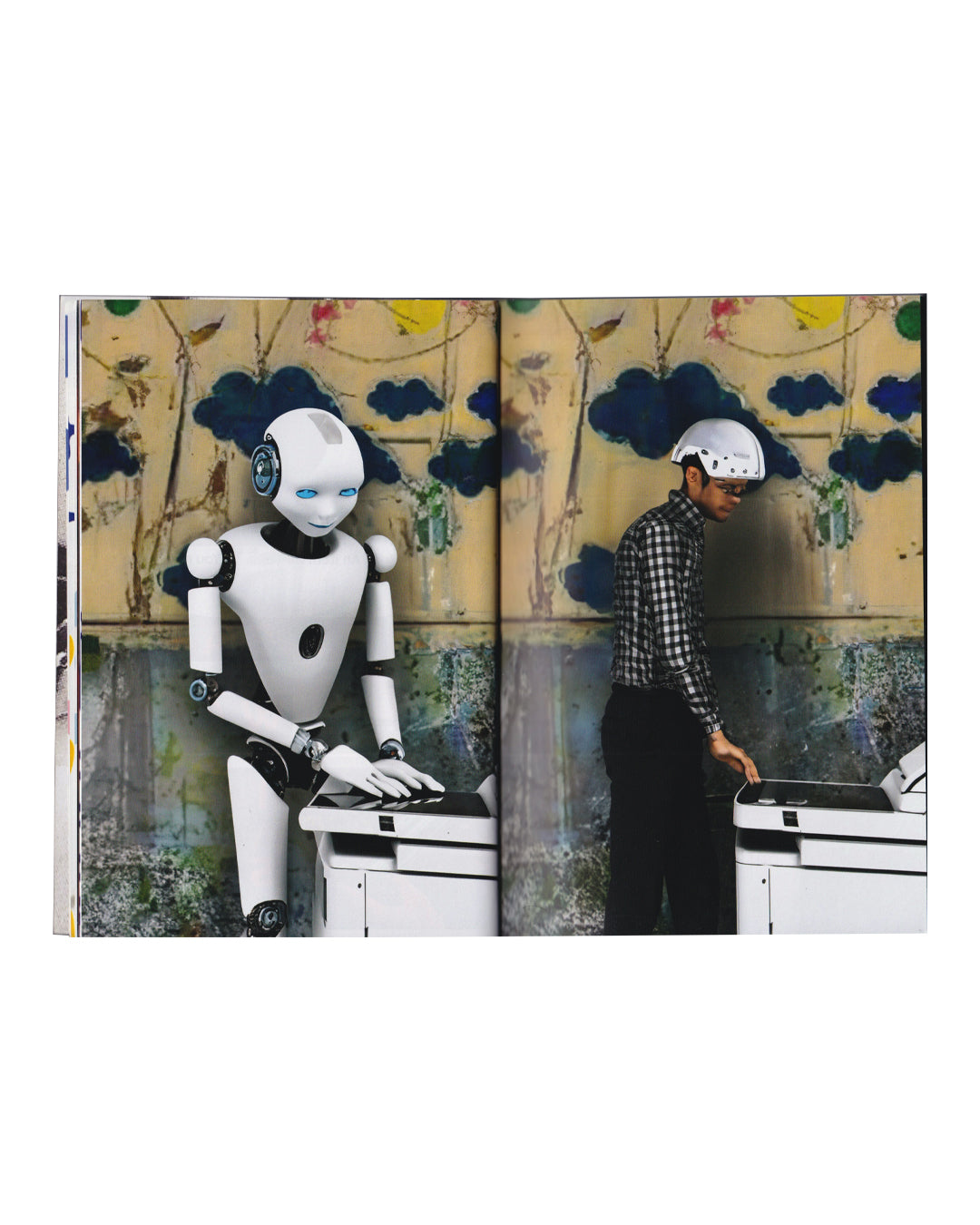 Rafael Melendez, The Naked Lunch, 2023 — interior spread; continuous full-bleed colour photograph across both pages showing a white humanoid robot with blue eyes seated at a white machine on the left, and a man in a checked shirt and white helmet operating an identical machine on the right; decorative painted wall with cloud and tree motifs behind both figures; robot and human in mirrored posture