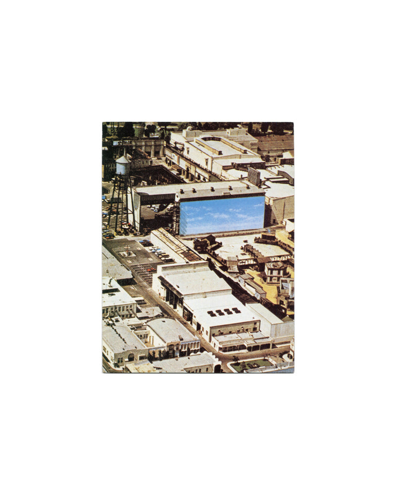 Exterior front of 1984 Alibis Centre Georges Pompidou exhibition invitation card, folded, showing a colour aerial photograph of a film studio backlot with a large rectangular billboard painted with a flat blue sky at centre, water tower at upper left, and surrounding low-rise industrial rooftops, printed to the right half of the folded card on semi-gloss stock.