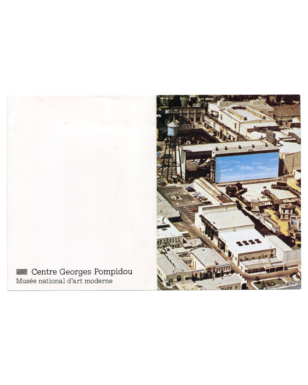 Exterior of 1984 Alibis Centre Georges Pompidou exhibition invitation card unfolded, showing right-hand panel with colour aerial photograph of a film studio backlot: low-rise industrial buildings, a water tower at left, and a large rectangular billboard structure painted with a flat blue sky, dominating the centre of the composition; left panel white with Centre Georges Pompidou / Musée national d'art moderne logotype at lower left in small black sans-serif type.