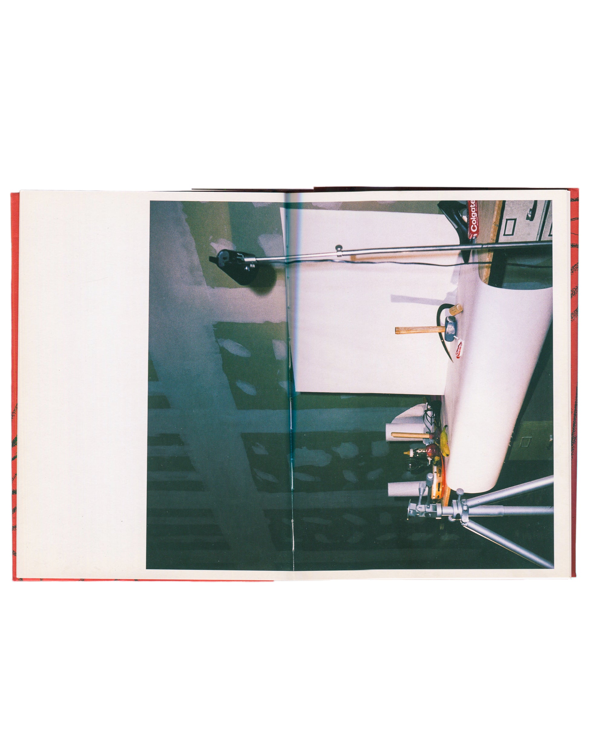 Interior spread, Andy Warhol, Hammer and Sickle, Haunch of Venison, London, 2004. Left page: blank white. Right page: full-bleed colour photograph — overhead view of Warhol's studio floor; large roll of white paper, camera on tripod, lighting equipment, 'Colgate' branded box visible upper right; dark green floor with shadow patterns. Red cloth binding visible at left edge.