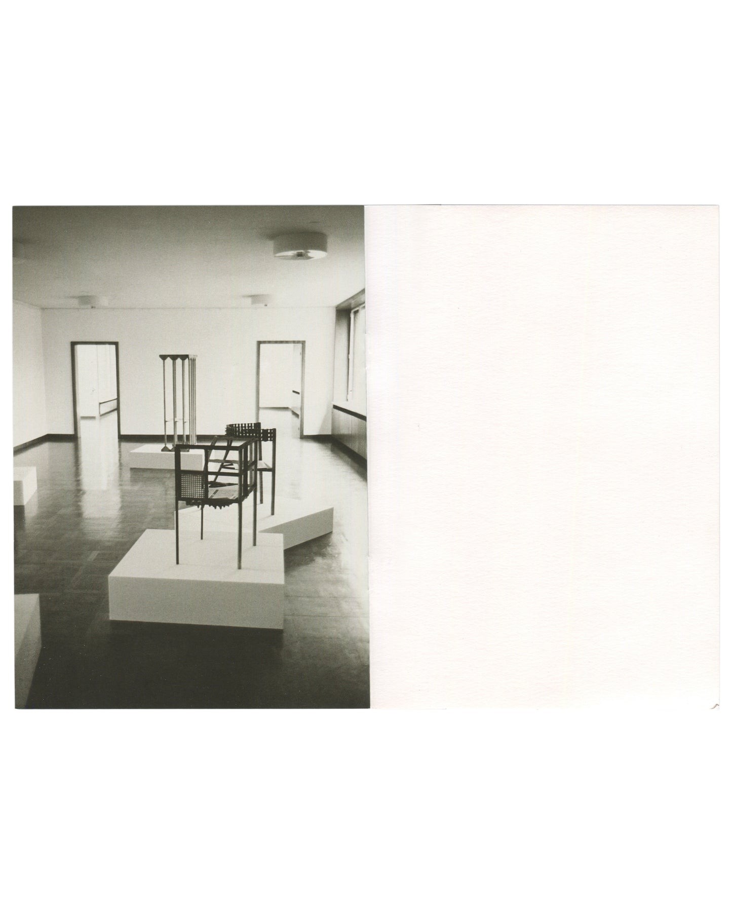 Interior spread, Franz West, Museum Haus Lange, Heft 2, Krefelder Kunstmuseen, Krefeld, 1989. Black-and-white installation photograph by Günther Förg — wire-frame and perforated-metal chair sculpture on white plinth foreground; tall multi-legged stool form on plinth behind; dark reflective floor, white walls, two doorways visible. Right page: blank white.