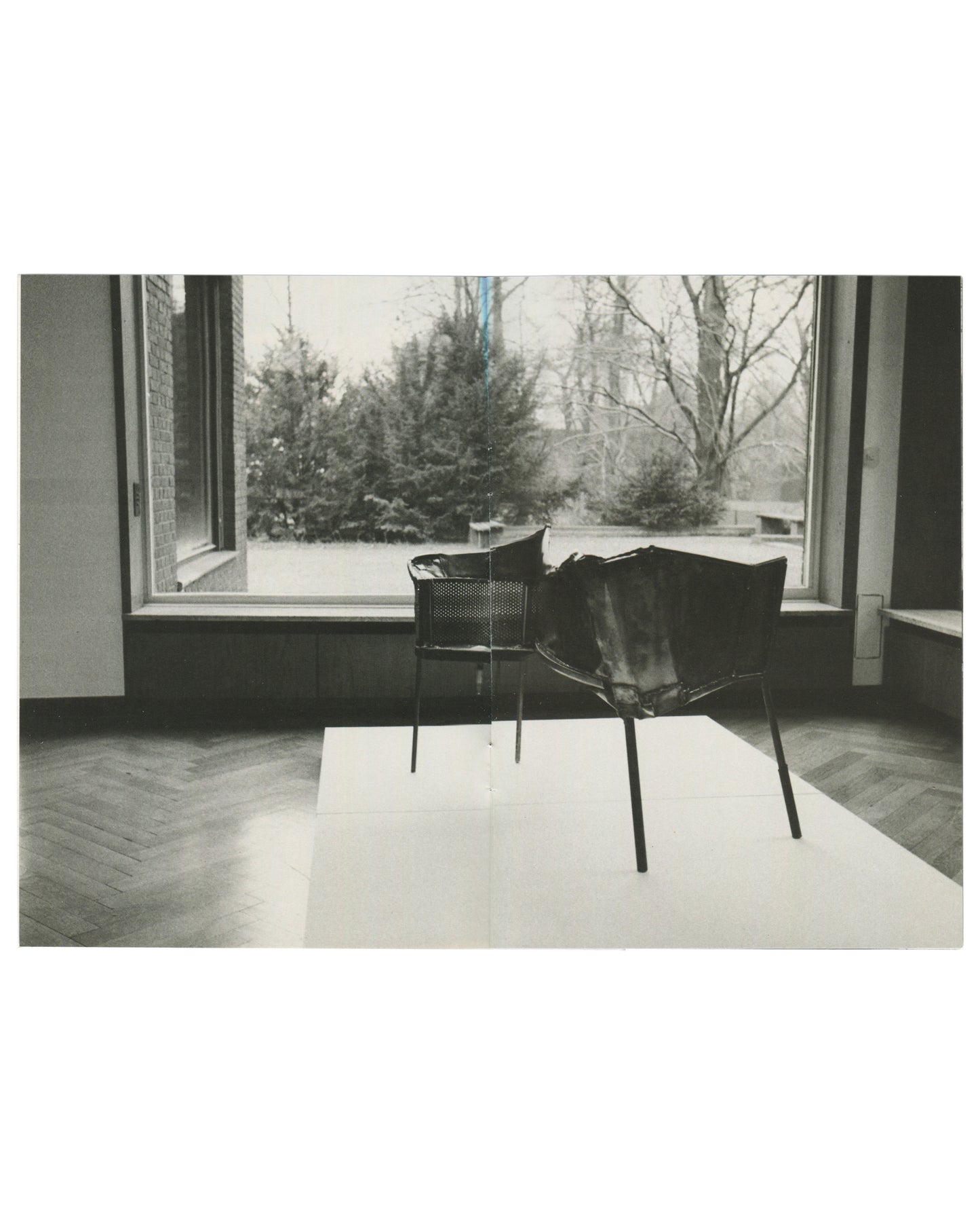 Interior spread, Franz West, Museum Haus Lange, Heft 2, Krefelder Kunstmuseen, Krefeld, 1989. Black-and-white photograph by Günther Förg — two sculptural chair forms on a white rug before a large window; perforated metal basket-seat left, draped leather or fabric form on metal frame right; winter garden with evergreens and bare trees outside; herringbone parquet floor.