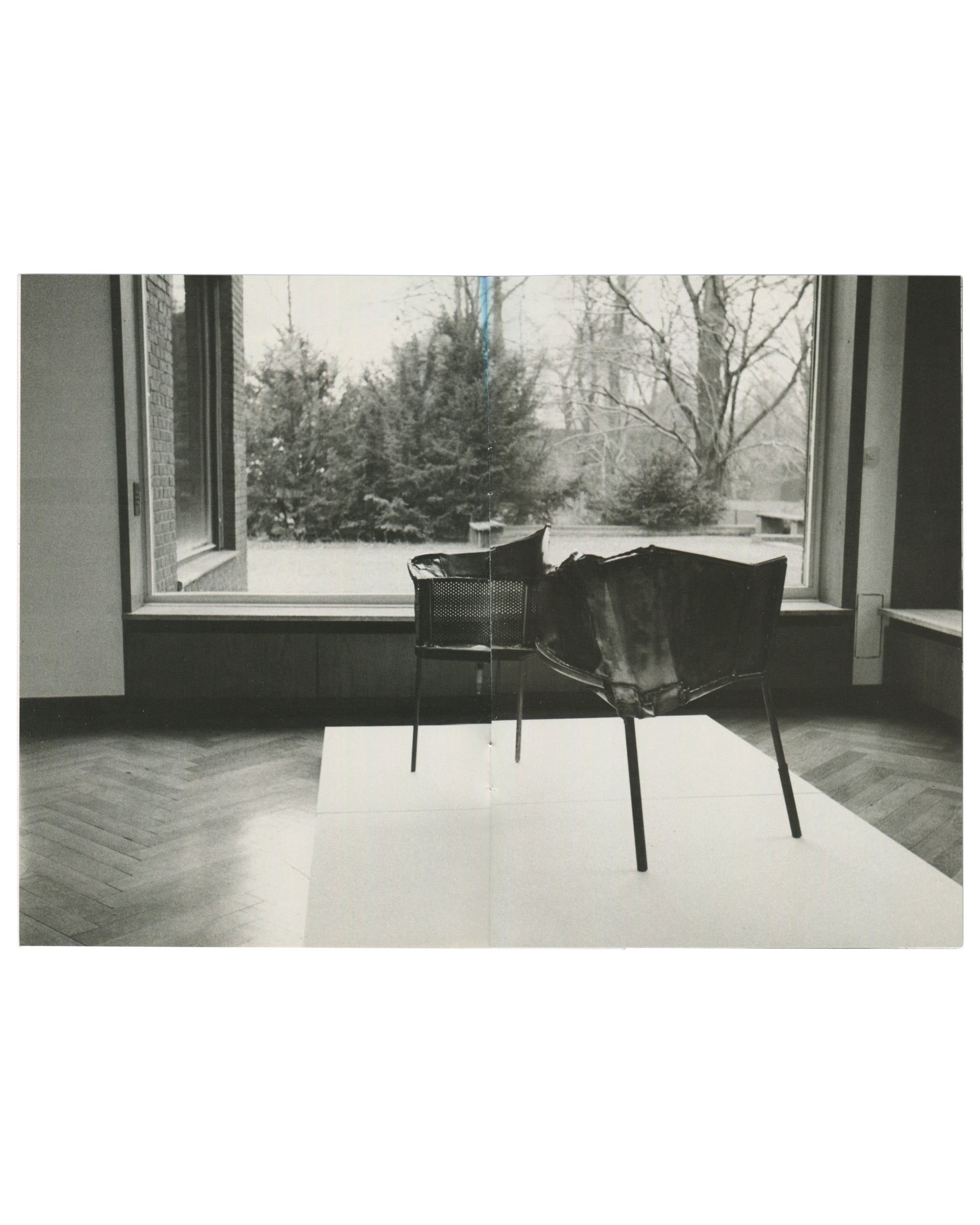Interior spread, Franz West, Museum Haus Lange, Heft 2, Krefelder Kunstmuseen, Krefeld, 1989. Black-and-white photograph by Günther Förg — two sculptural chair forms on a white rug before a large window; perforated metal basket-seat left, draped leather or fabric form on metal frame right; winter garden with evergreens and bare trees outside; herringbone parquet floor.