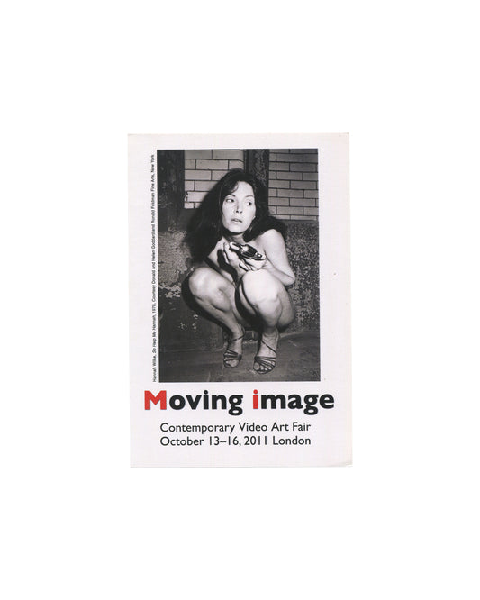 Front Moving Image Contemporary Video Art Fair, London, 2011, announcement card, front, white card: black-and-white photograph of Hannah Wilke crouching against a brick wall, holding a shoe, from So Help Me Hannah, 1978; Moving image in large type below with M and i in red; Contemporary Video Art Fair / October 13–16, 2011 London in black sans-serif; vertical caption at left edge: Hannah Wilke, So Help Me Hannah, 1978. Courtesy Donald and Helen Goddard and Ronald Feldman Fine Arts, New York.
