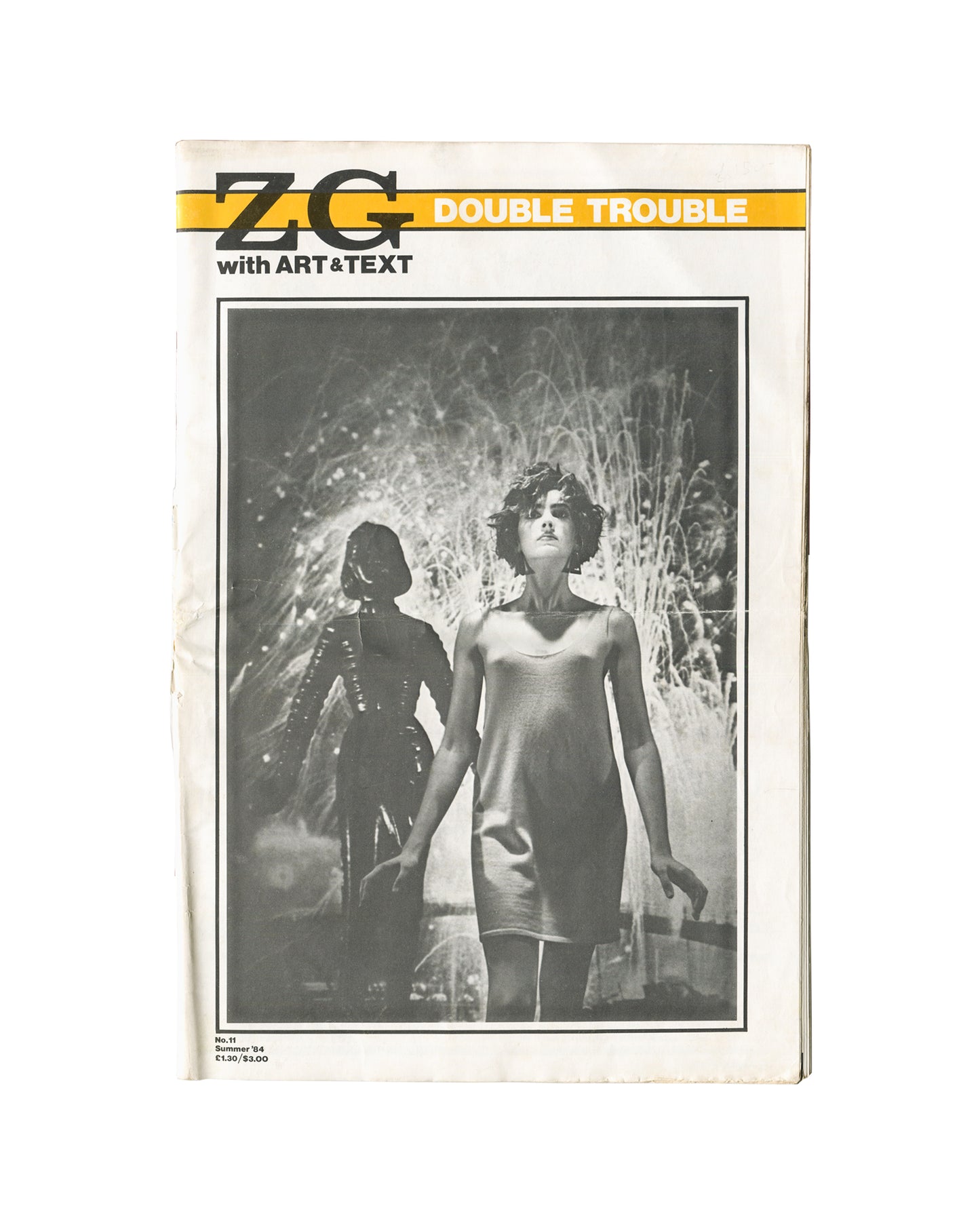 Magazine cover, ZG No. 11 / Art & Text 15: Double Trouble, ZG Magazine / Art & Text, New York / Melbourne, 1984. Edited by Rosetta Brooks. Large-format staple-bound periodical, 28 × 42 cm. Yellow horizontal band upper third — bold black 'ZG' logotype left; white sans-serif 'DOUBLE TROUBLE' right; below band: 'with ART&TEXT'. Full-bleed black-and-white photograph — two female figures in hazy backlit environment; foreground figure in short slip dress, short dark hair; background figure in dark structured outf
