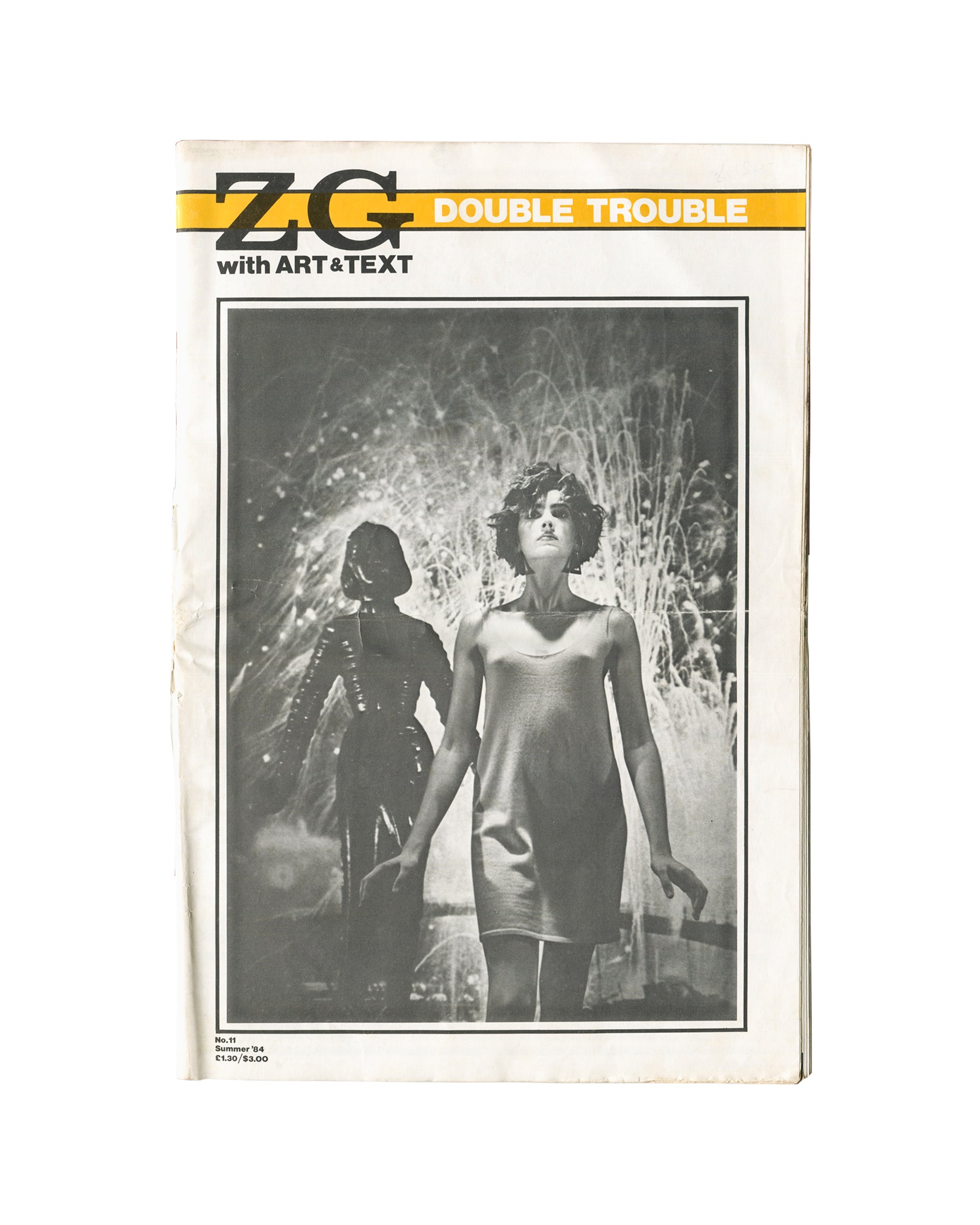 Magazine cover, ZG No. 11 / Art & Text 15: Double Trouble, ZG Magazine / Art & Text, New York / Melbourne, 1984. Edited by Rosetta Brooks. Large-format staple-bound periodical, 28 × 42 cm. Yellow horizontal band upper third — bold black 'ZG' logotype left; white sans-serif 'DOUBLE TROUBLE' right; below band: 'with ART&TEXT'. Full-bleed black-and-white photograph — two female figures in hazy backlit environment; foreground figure in short slip dress, short dark hair; background figure in dark structured outf